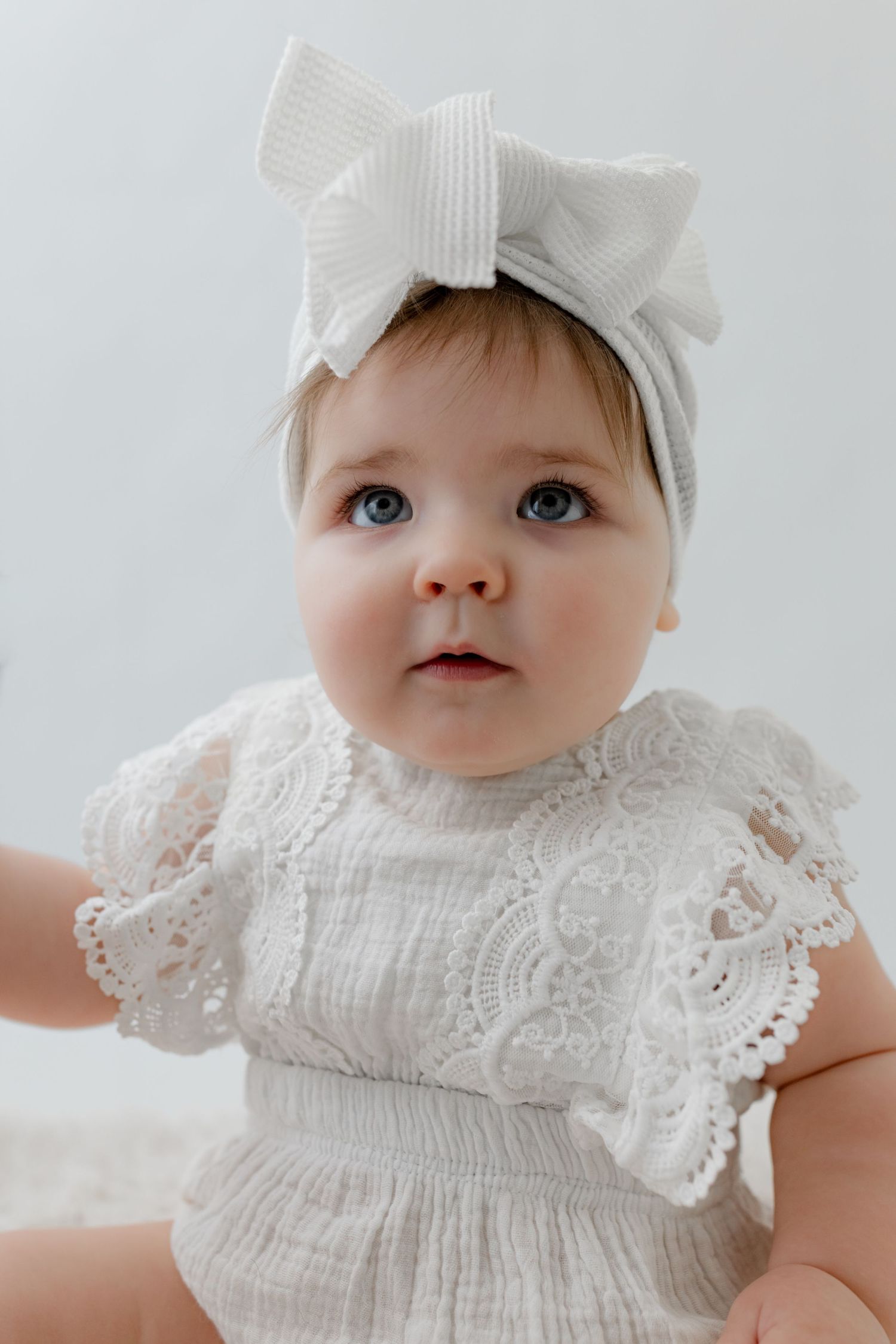 Baby girl in white lace romper and striped bow headband, with blue eyes, posed against a light background.