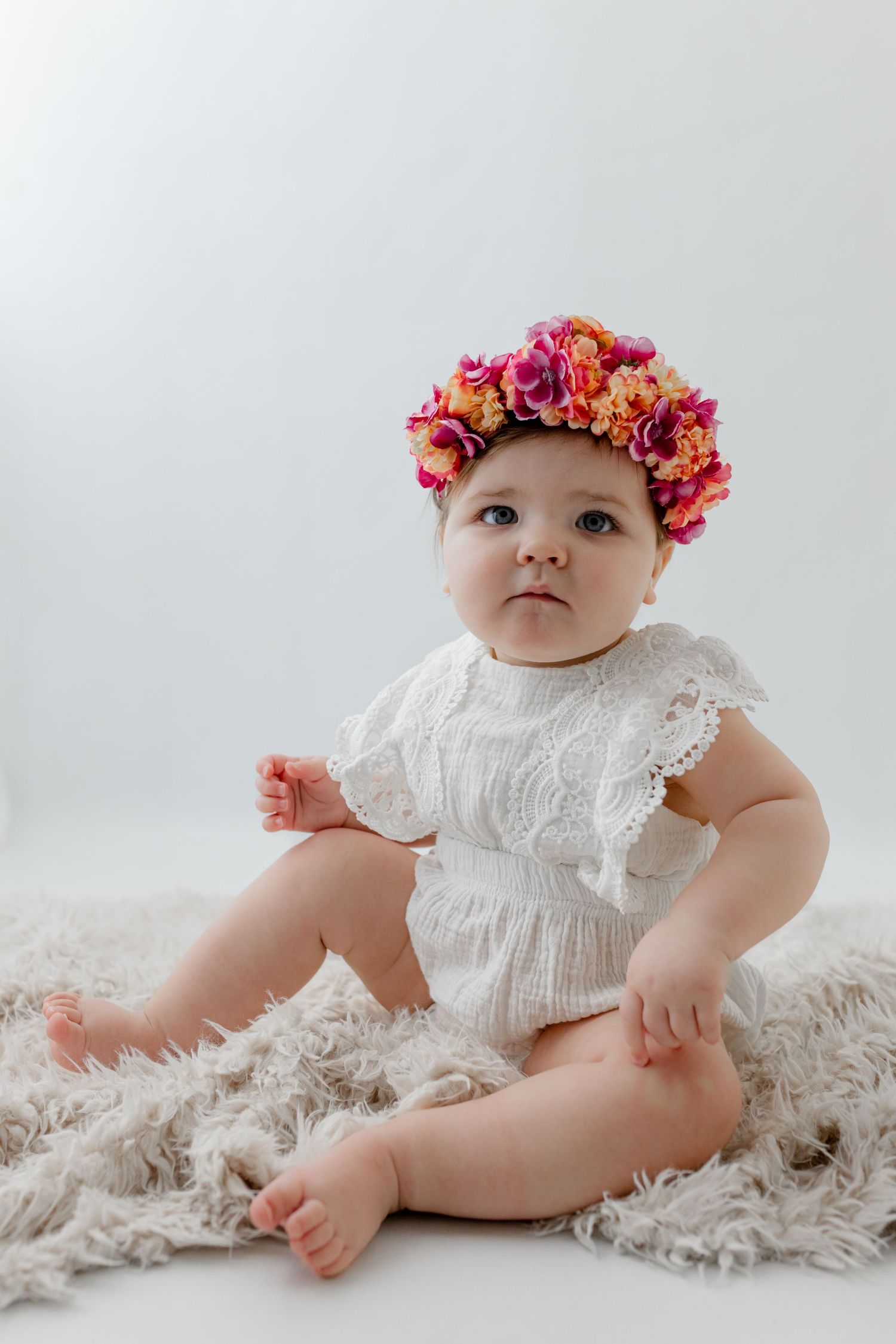 Baby girl in white lace romper and colorful floral crown sits on fluffy rug in bright studio photo.