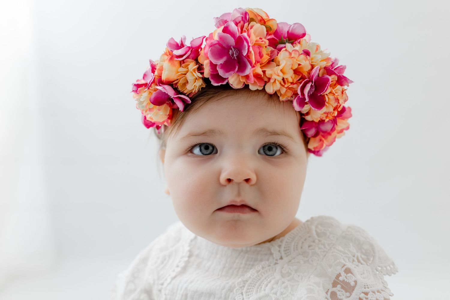 Baby girl wearing a colorful pink and orange floral crown headband, dressed in white lace, with blue eyes.