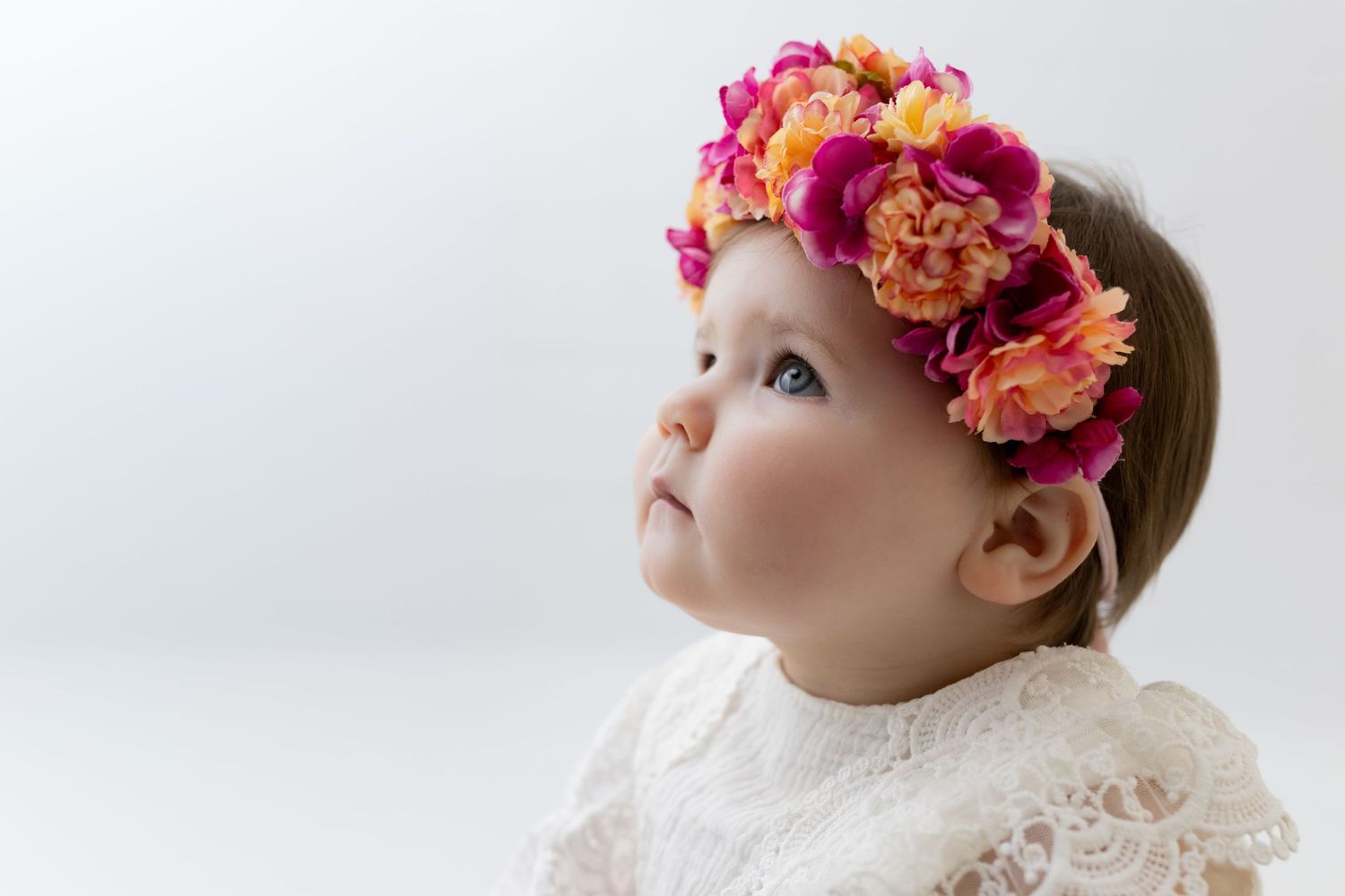 Baby girl with blue eyes wearing a pink and orange floral crown headband, looking up against white background.