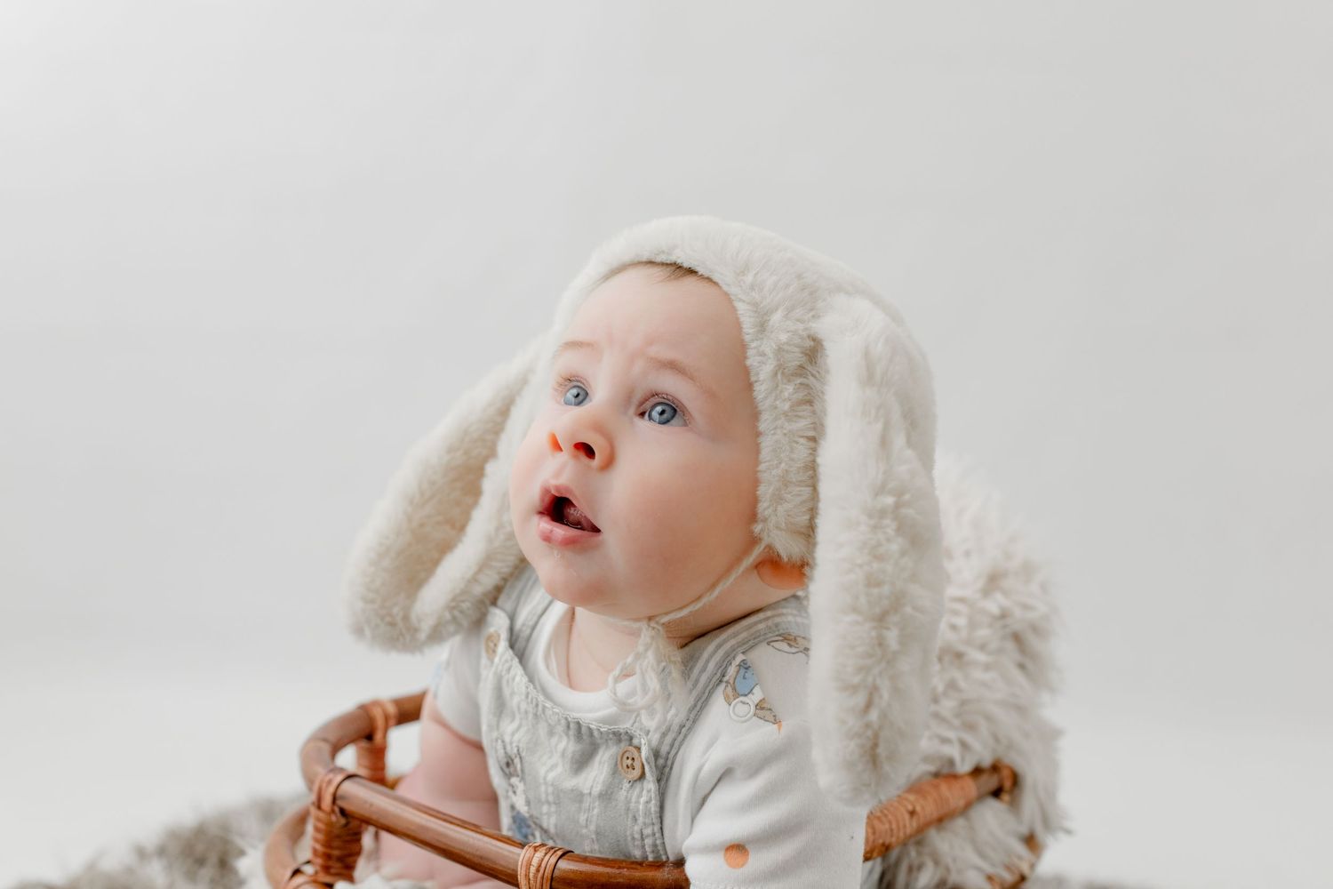 Adorable baby with blue eyes wearing a fluffy bunny ear hat sits in a wicker basket against a white background.