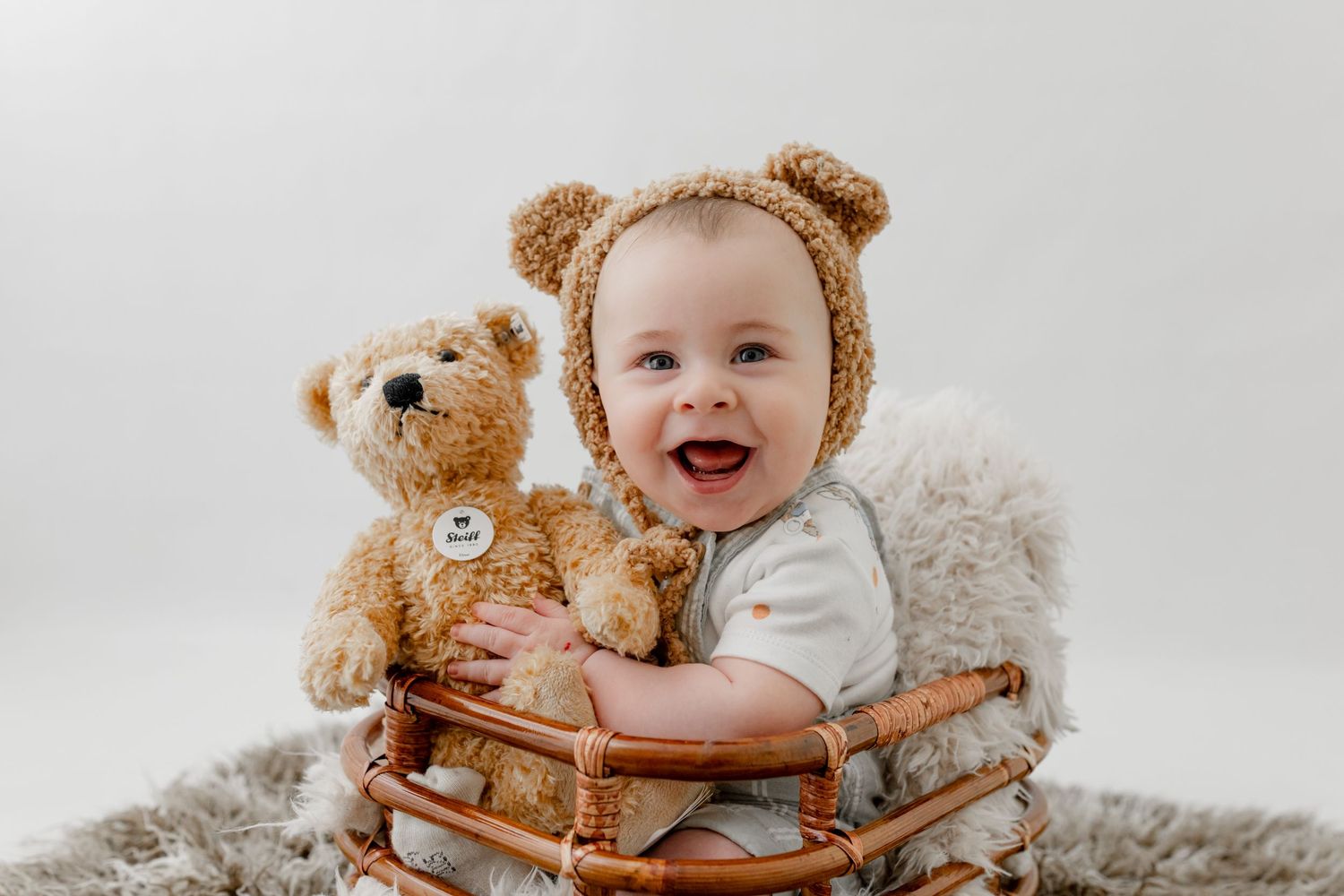 Smiling baby in bear bonnet sits in wicker basket hugging a teddy bear on a white background.