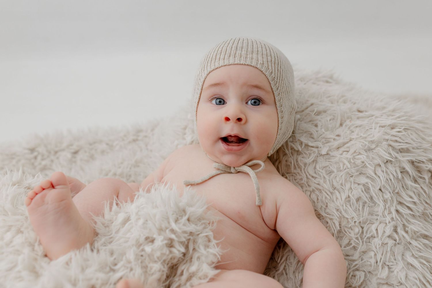Adorable baby wearing a beige knit bonnet lying on a fluffy white blanket with bright blue eyes and an open mouth smile.