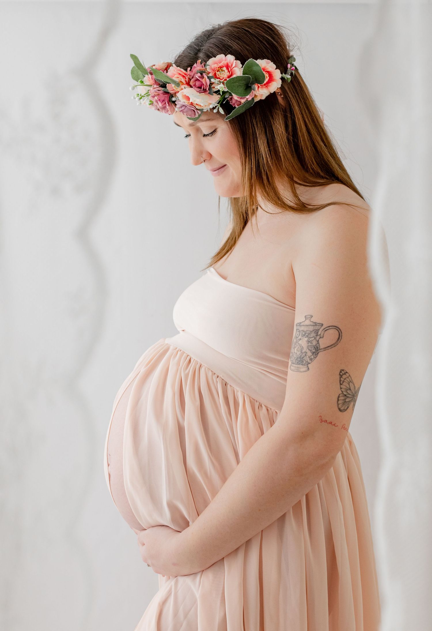 Pregnant woman in blush strapless gown and floral crown cradling baby bump, side profile maternity portrait.