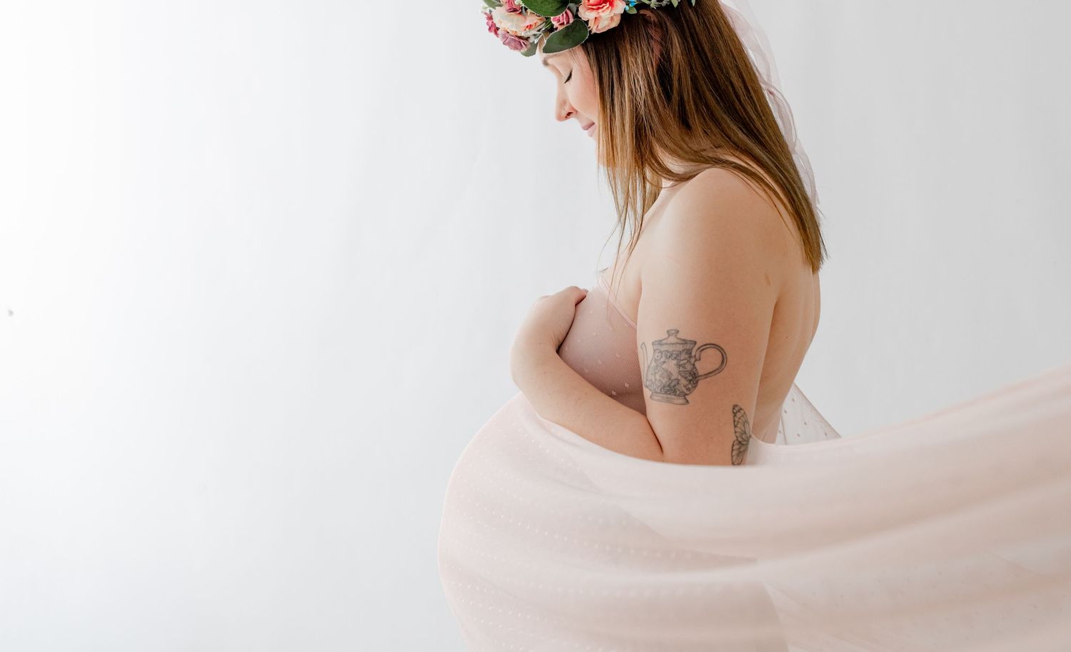 Pregnant woman in floral crown and sheer pink fabric wrap poses gracefully in a soft white studio setting.
