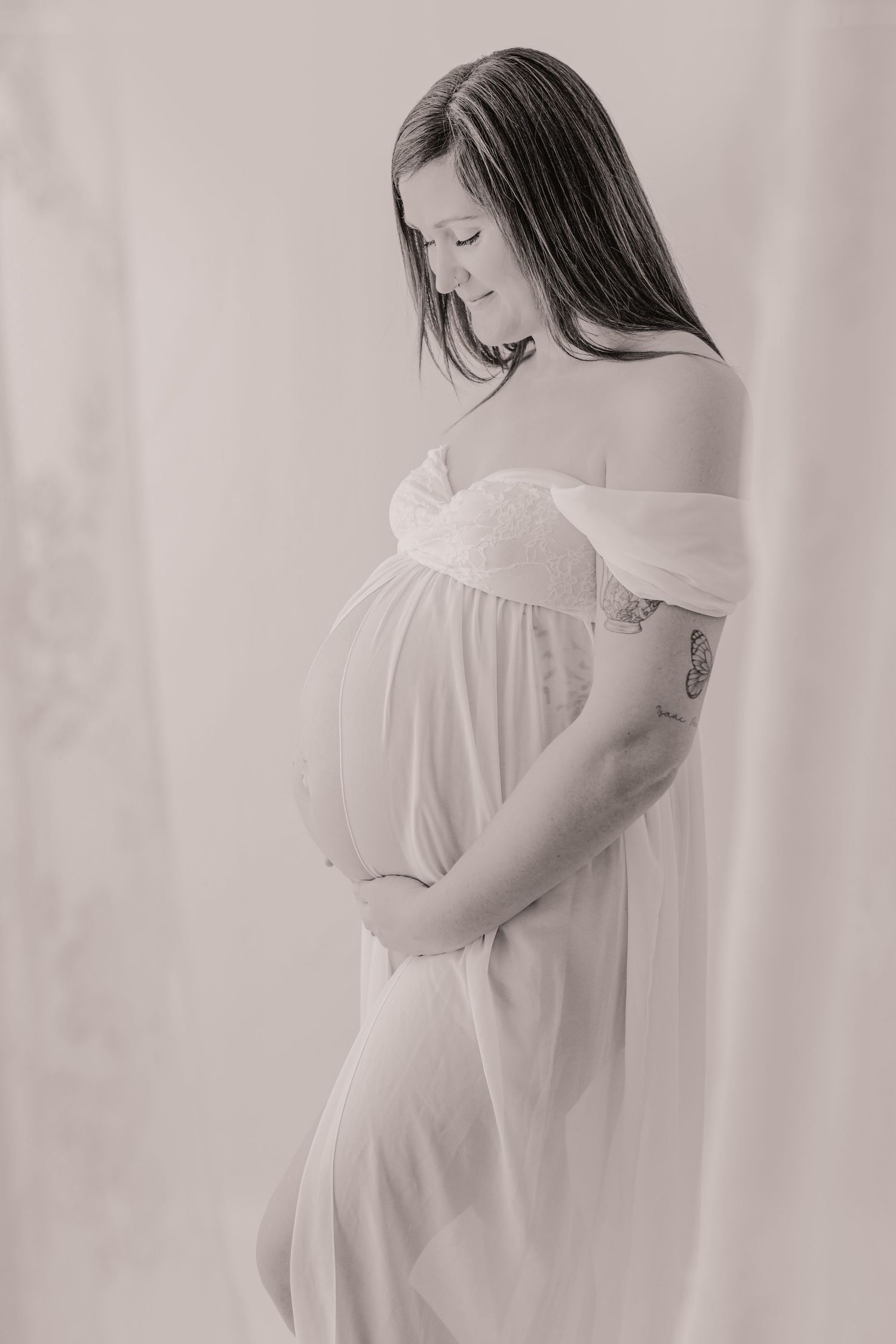 Elegant black and white maternity photo of a pregnant woman in a flowing off-shoulder white dress.