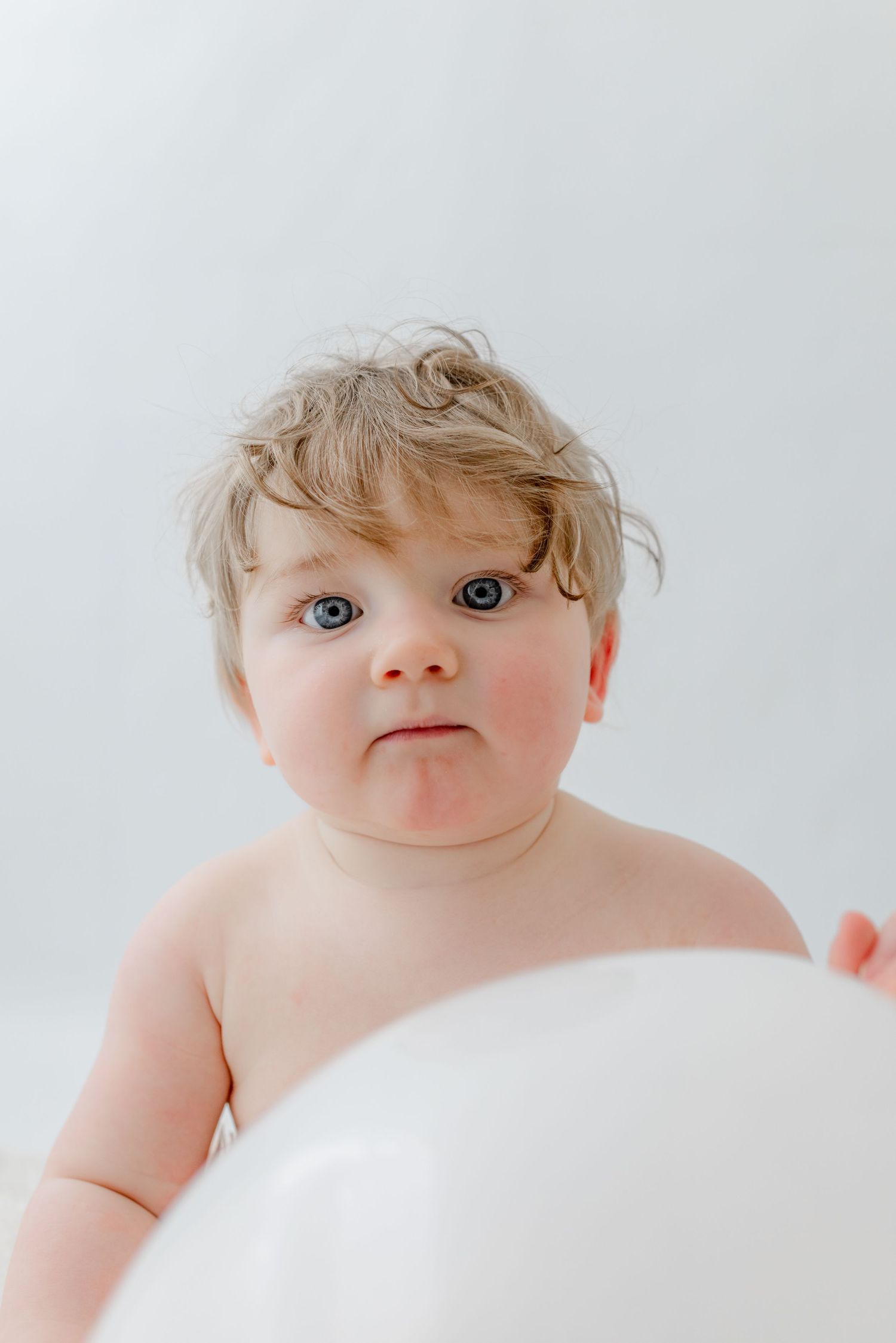 Cute blue-eyed baby with curly blonde hair making a funny face next to a white balloon.