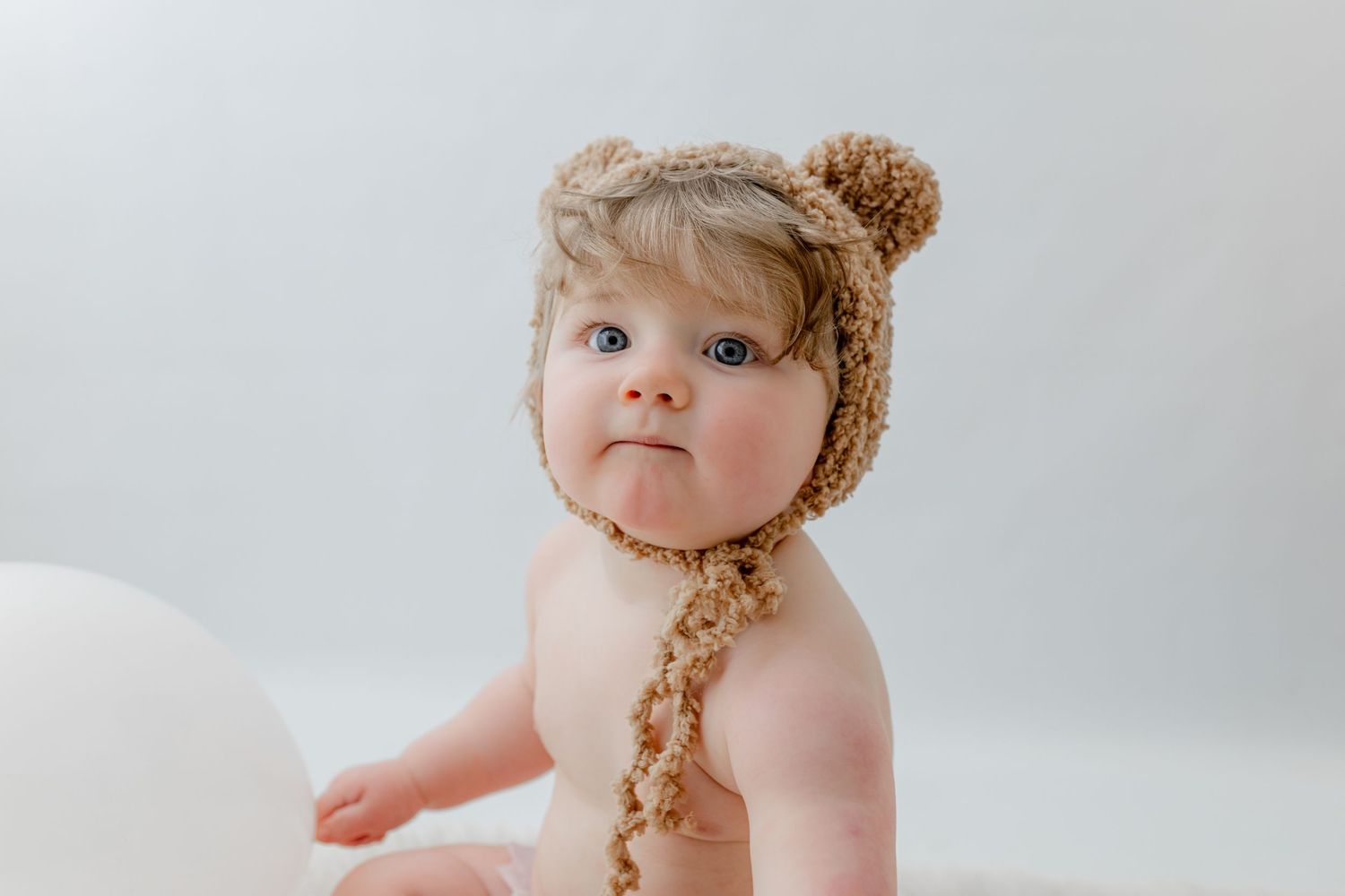 Adorable baby wearing a brown knit bear bonnet hat sitting against a white background.