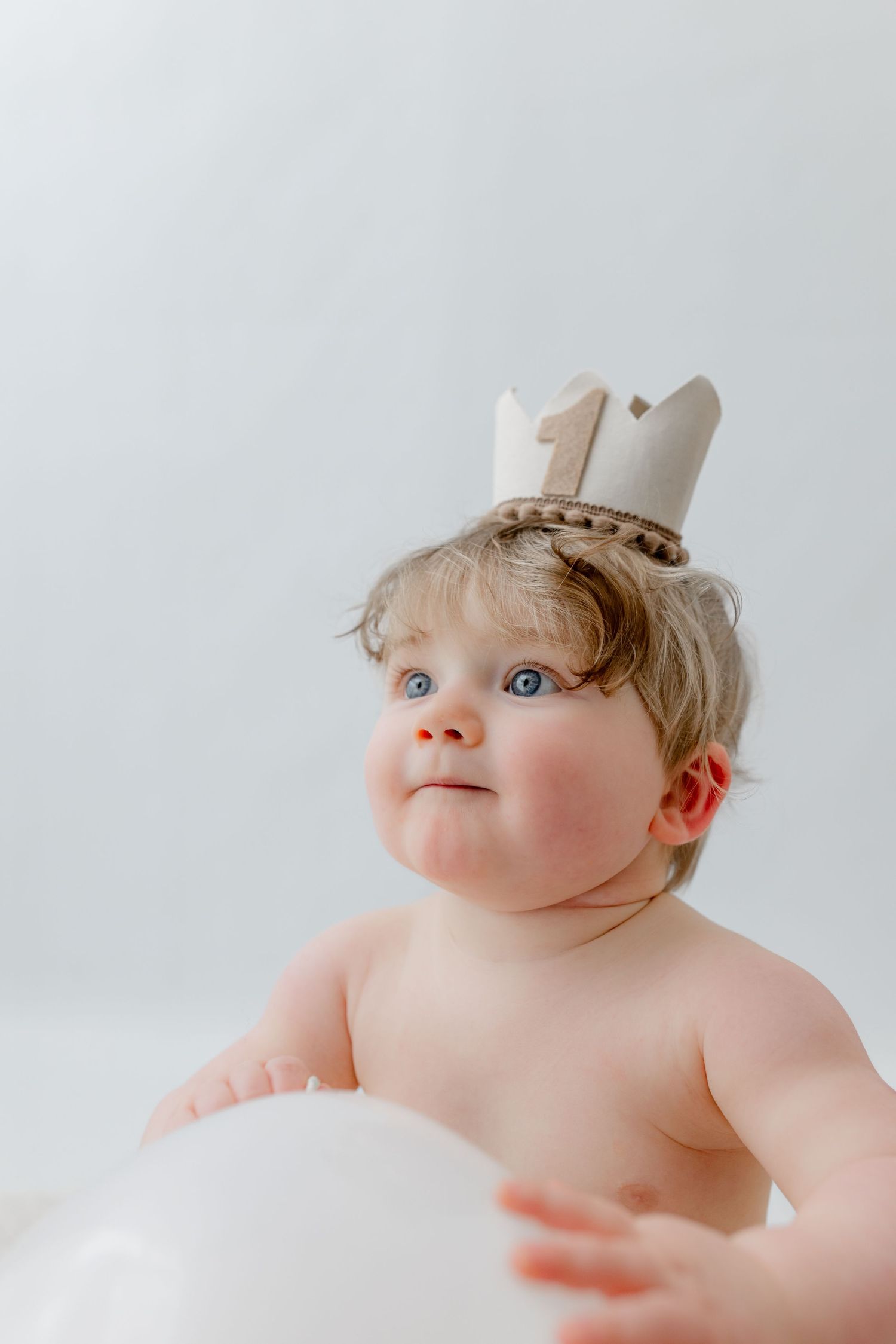 Adorable blue-eyed baby wearing a number one crown celebrating their first birthday with a white balloon.
