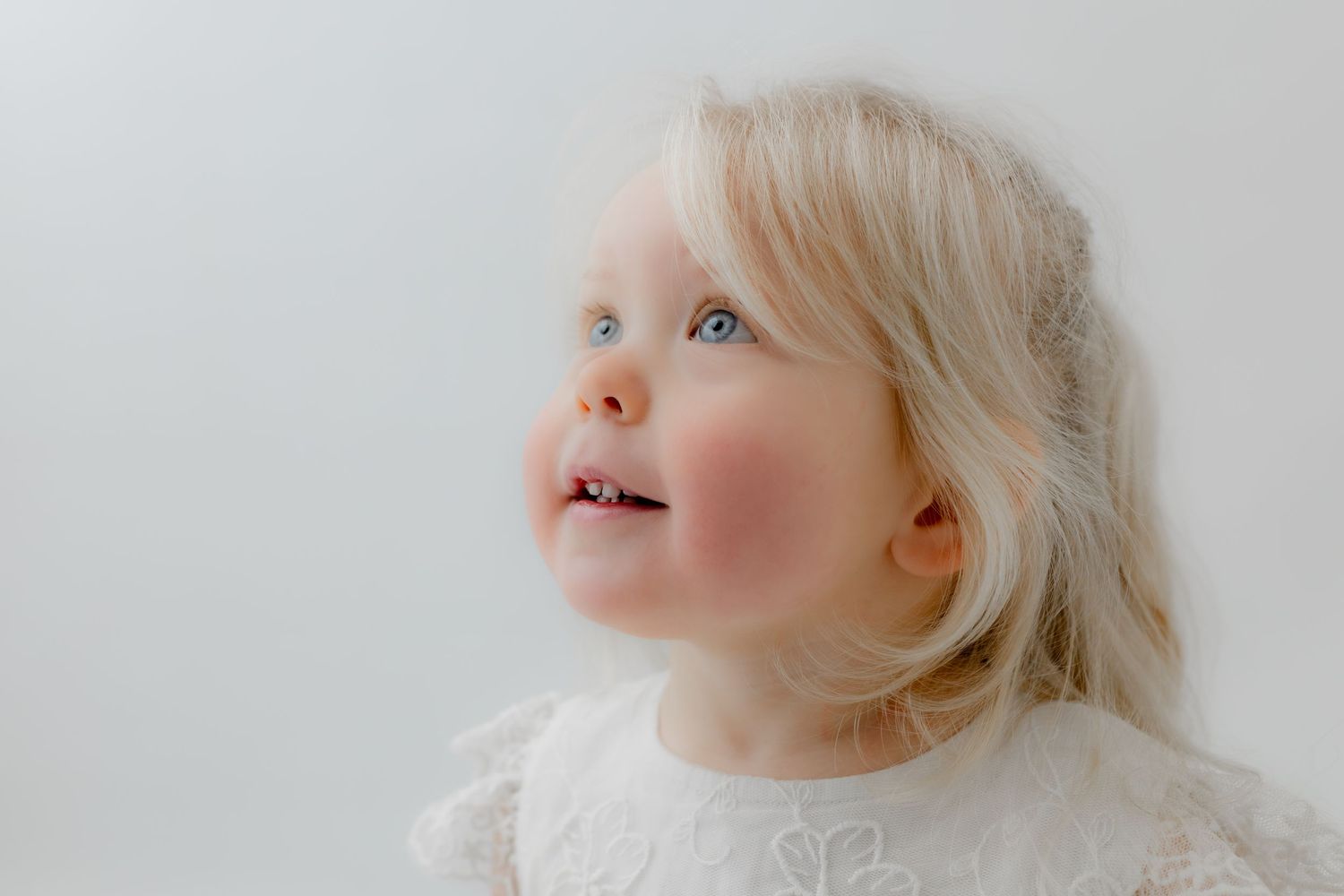 Adorable blonde toddler girl with blue eyes wearing a white lace dress, looking up with a sweet smile.