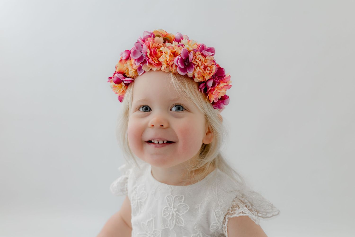 Smiling toddler girl wearing a vibrant pink and orange floral crown and white lace dress against a light background.