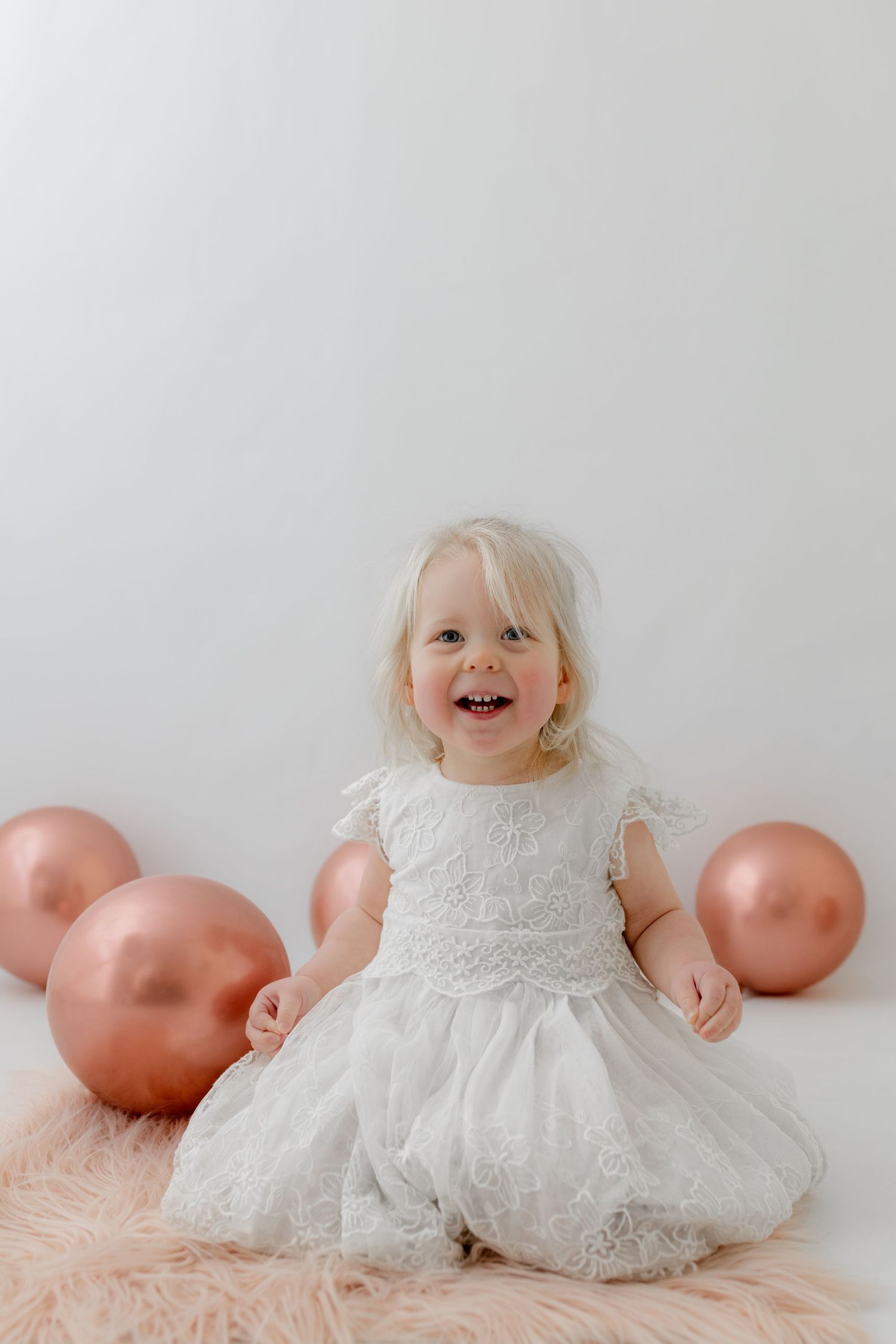 Smiling toddler girl in white lace dress sitting with rose gold balloons on a fluffy pink rug against white background.