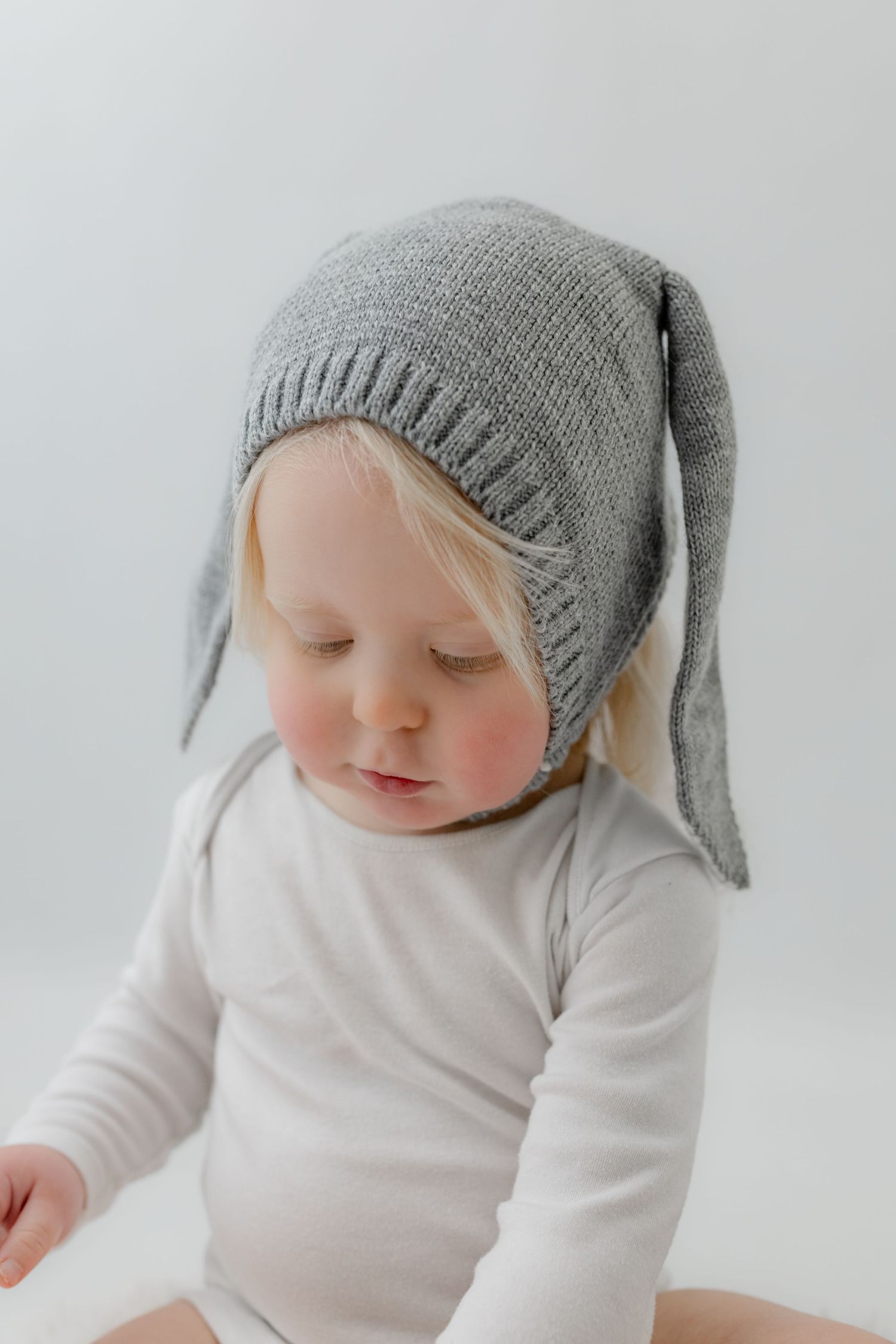 Baby wearing a gray knit bunny ear bonnet hat and white bodysuit, sitting against a light background.
