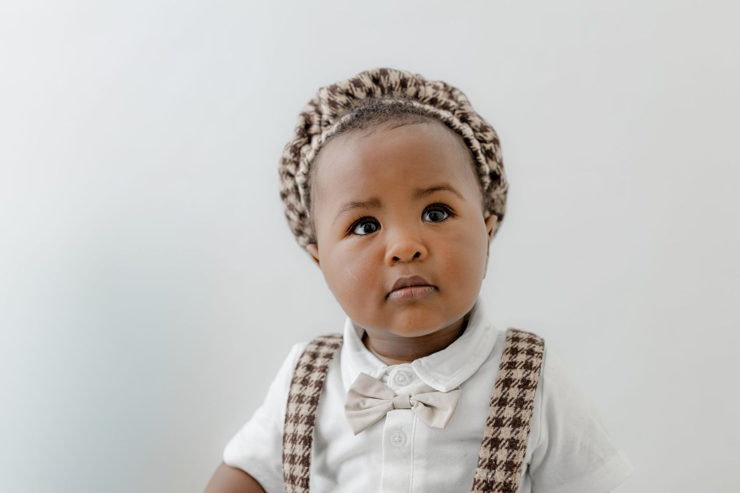 Adorable Black baby wearing a plaid beret hat and suspenders with a white bow tie shirt, posing against a white background.