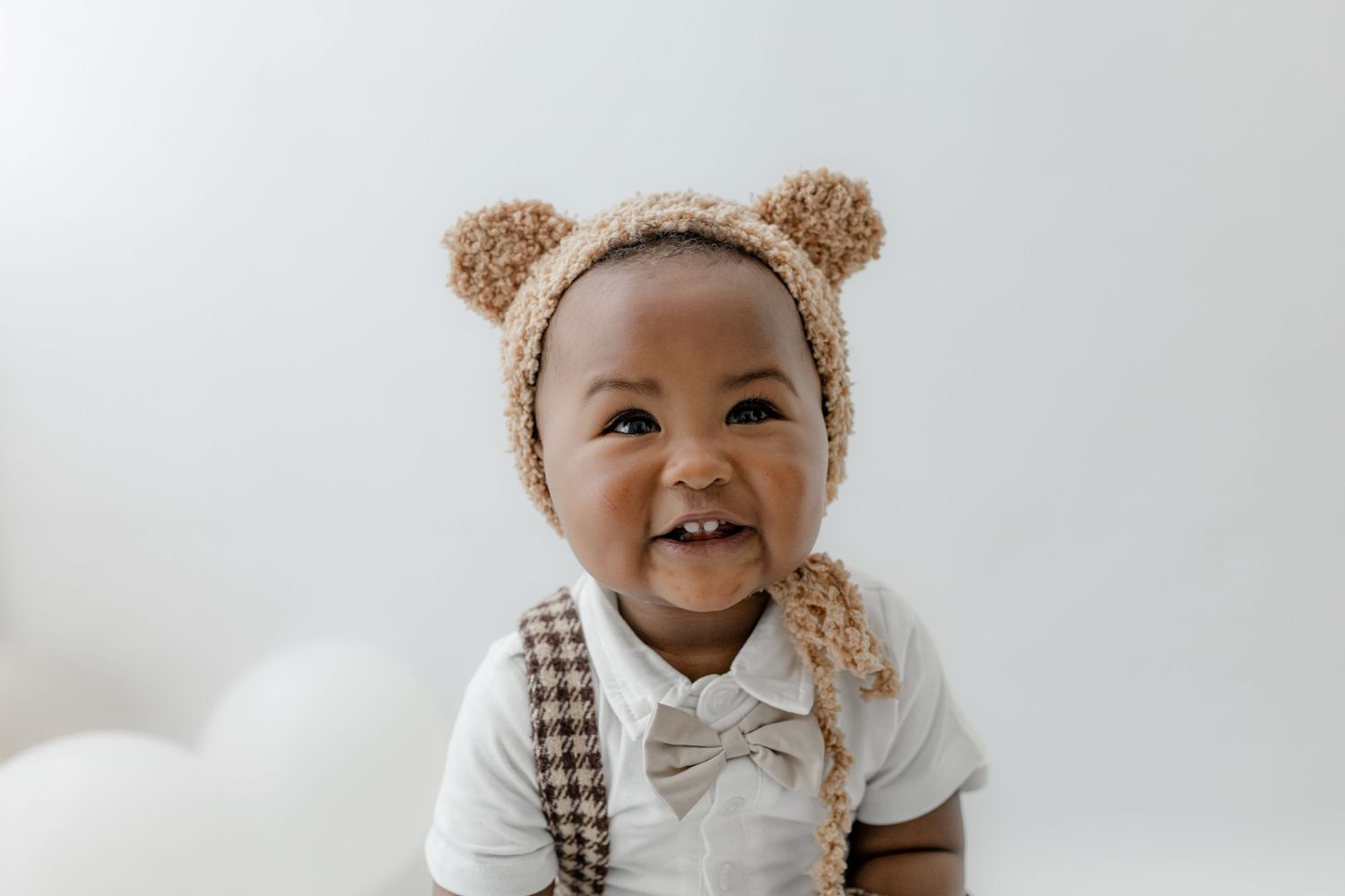 Smiling baby wearing a cute teddy bear bonnet and white outfit with bow tie against white background.