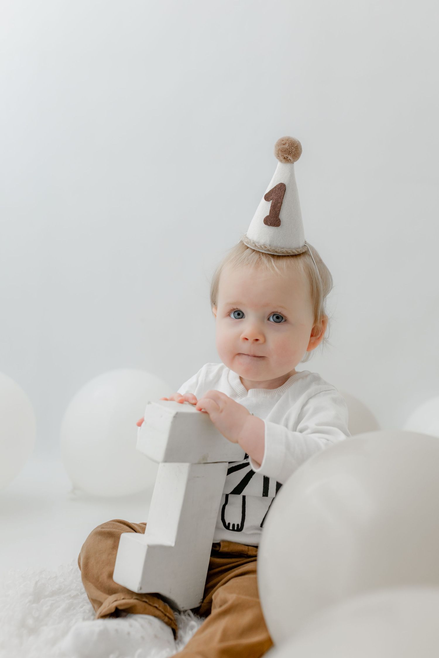 A baby wearing a number 1 birthday hat sits surrounded by white balloons, opening a gift box during their first birthday.