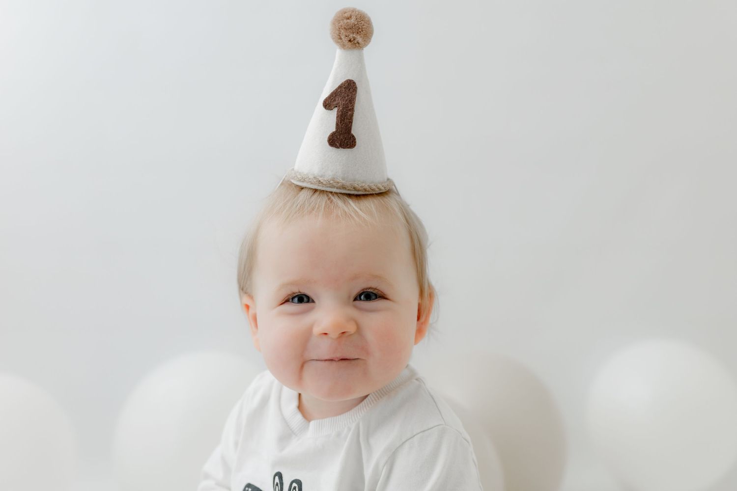 A smiling baby wearing a white number one birthday hat celebrates their first birthday surrounded by white balloons.