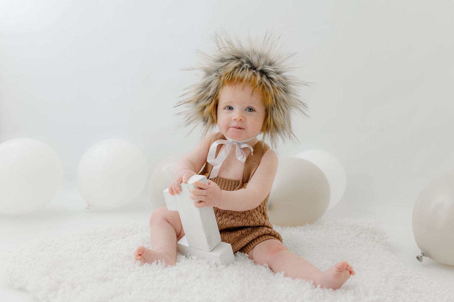 Baby in fuzzy hat and brown knit romper sitting on white fur rug with balloons holding a gift.