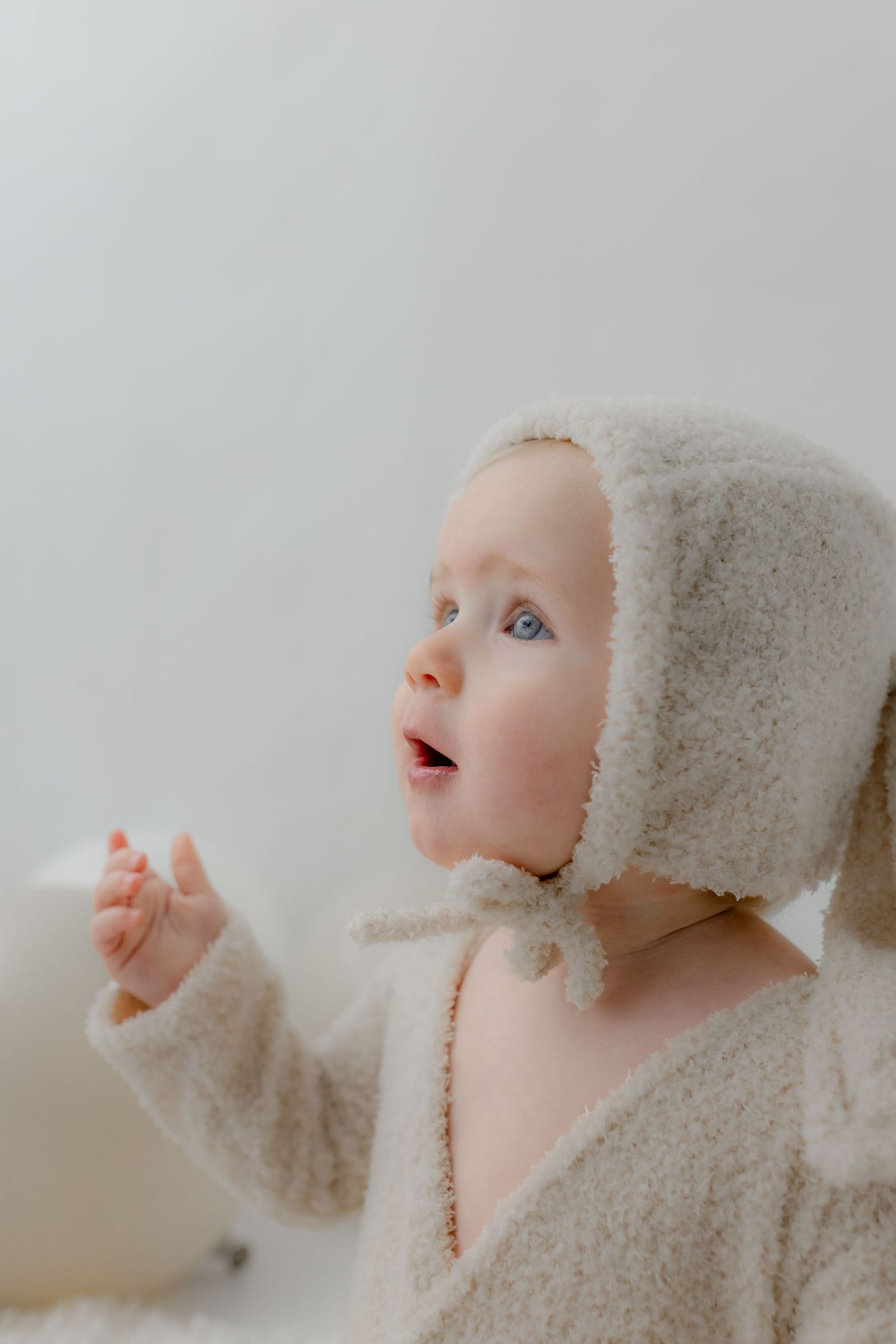 Adorable blue-eyed baby in a cozy cream sherpa bonnet and outfit, looking up with mouth open in wonder.