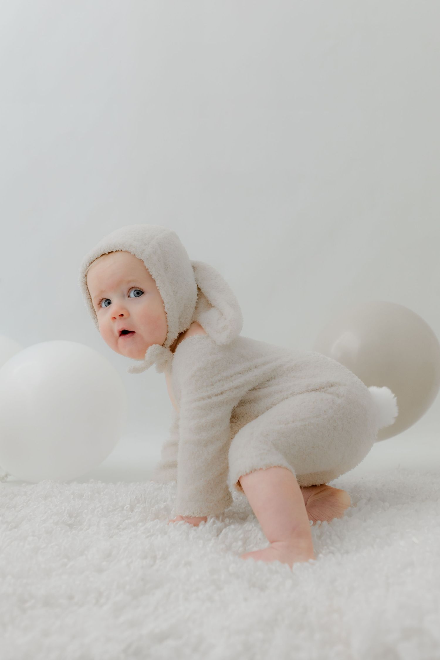 A baby in a cream fuzzy lamb costume crawls on a white fluffy carpet surrounded by white balloons.