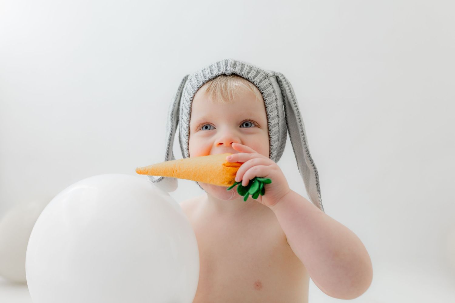Baby wearing gray bunny ears hat chewing on a carrot toy next to a white balloon on white background.