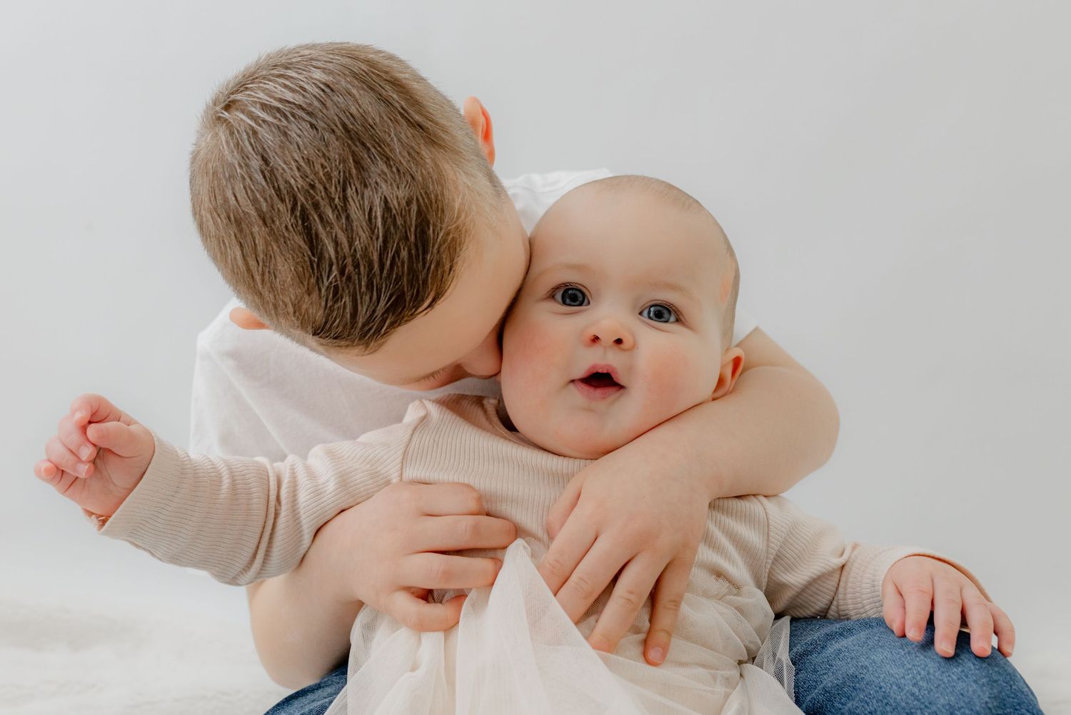 An older toddler boy hugs and kisses a wide-eyed baby sibling against a clean white background.