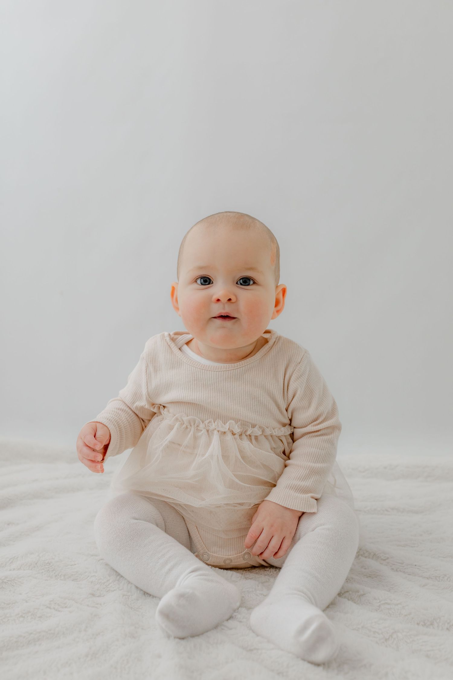 A cute baby in a beige long-sleeve romper and white tights sits on a soft white blanket against a light gray background.