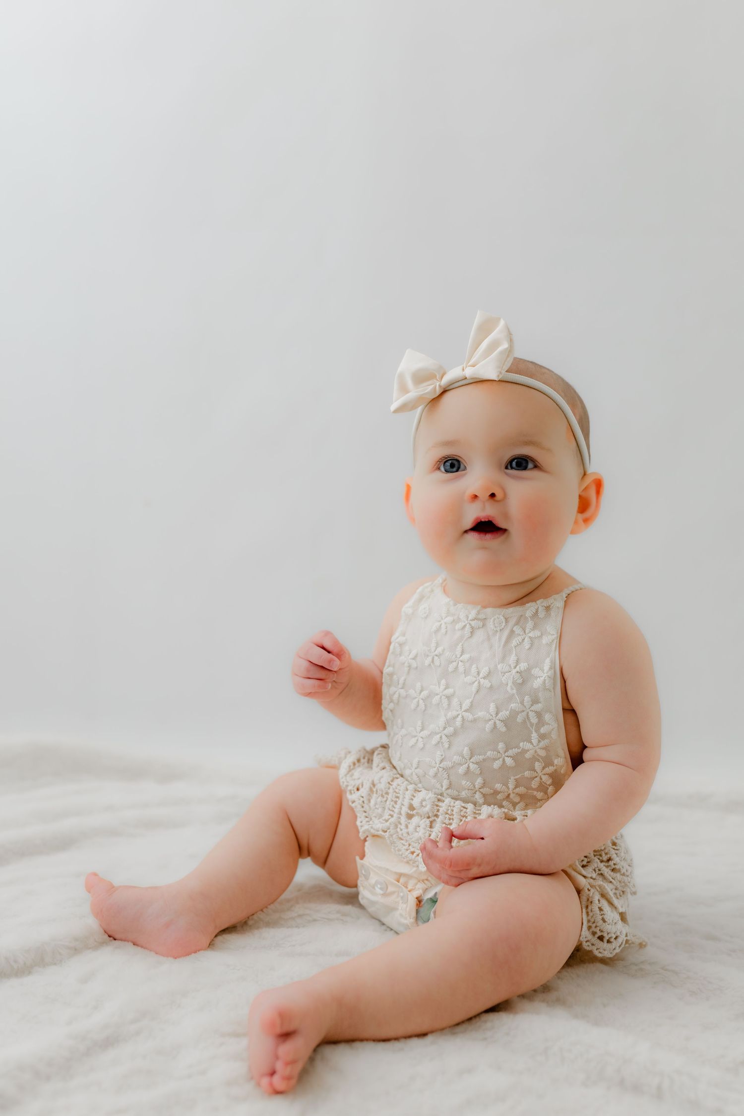 Adorable baby girl in cream lace romper and bow headband sitting on white blanket, looking up curiously.