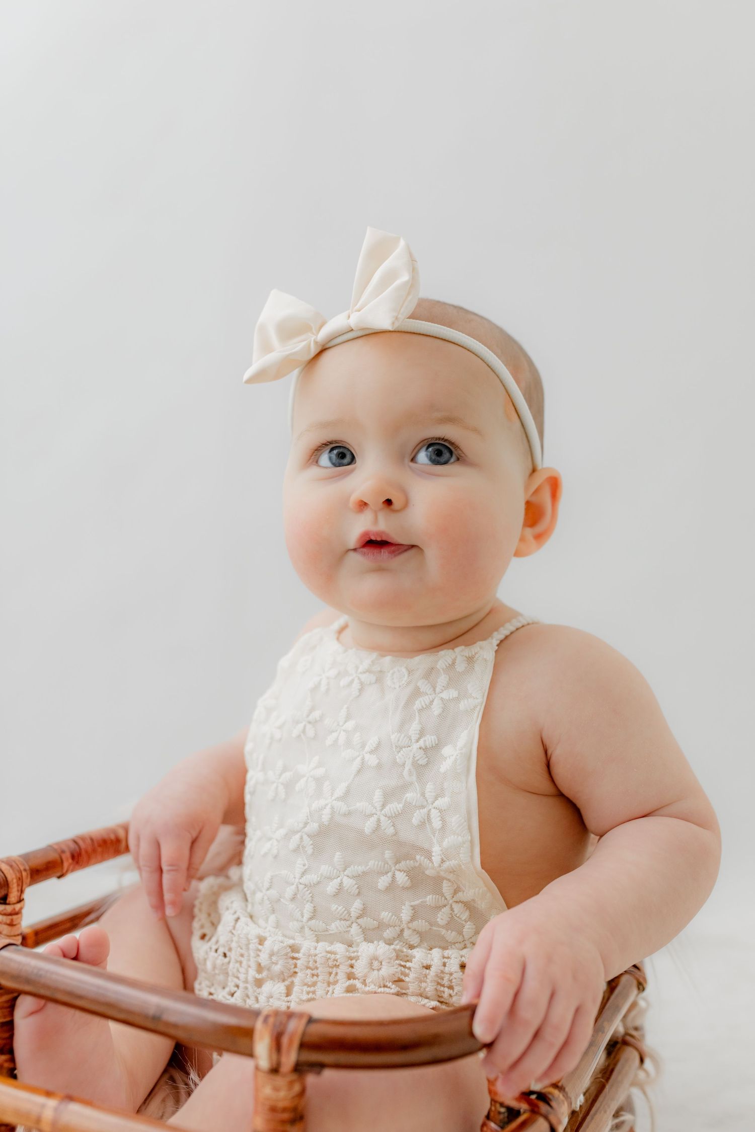 Adorable baby girl with blue eyes wearing a white floral romper and cream bow headband sitting in a wicker chair.