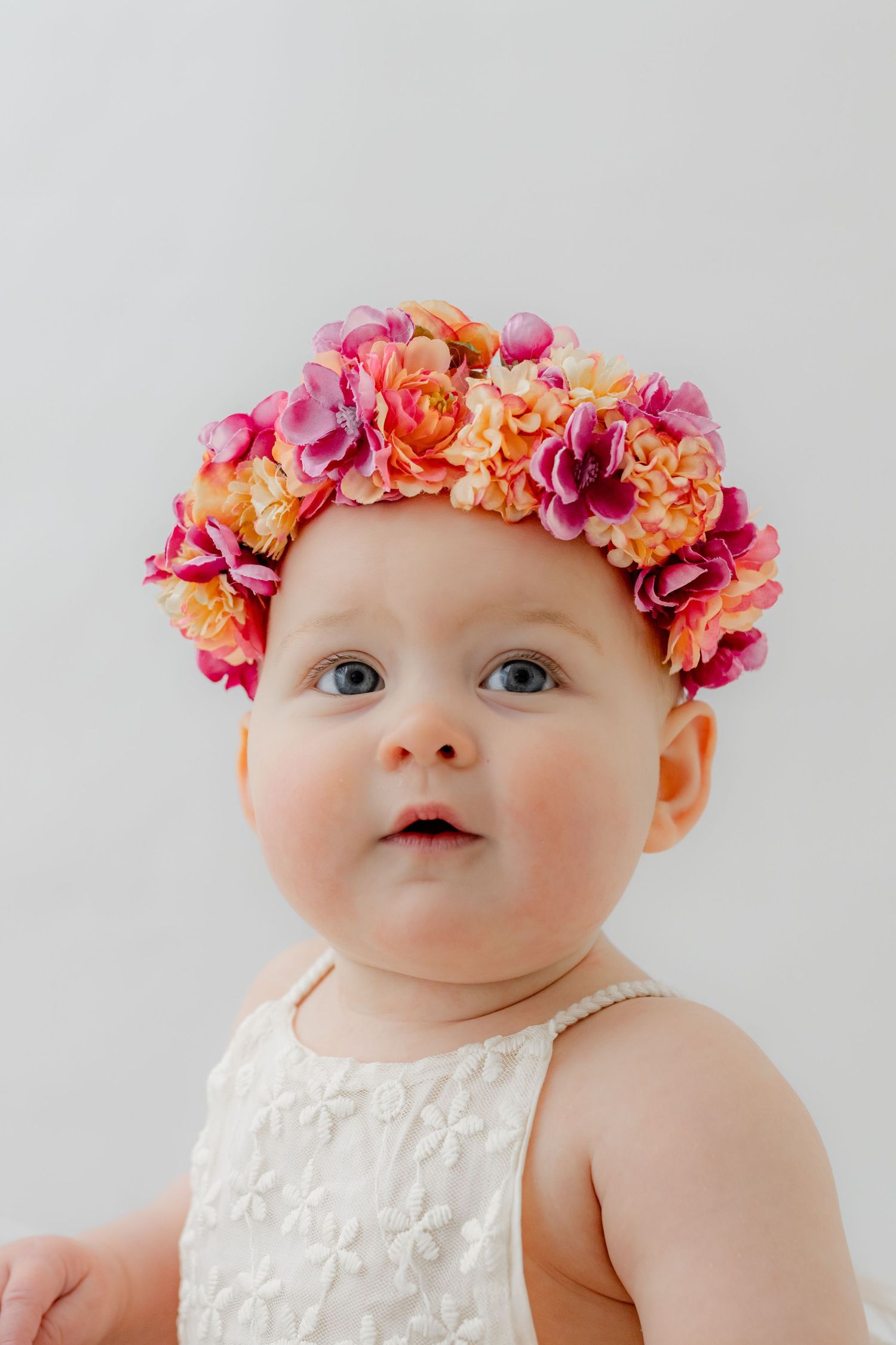 Adorable baby girl wearing a colorful pink and orange floral crown headband, dressed in white, with big blue eyes.