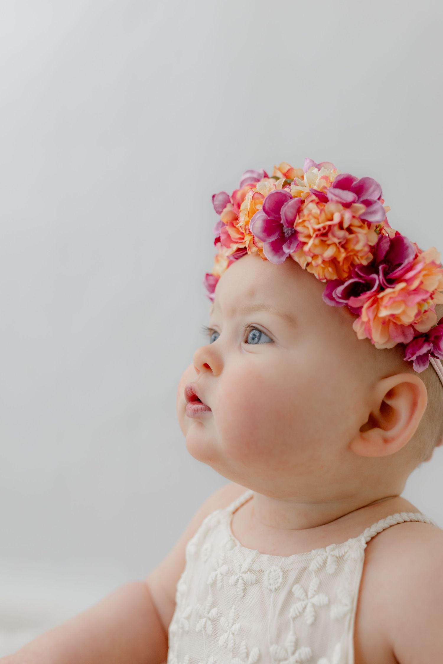 Baby girl wearing a colorful floral crown with pink and orange flowers, dressed in white, looking up curiously.