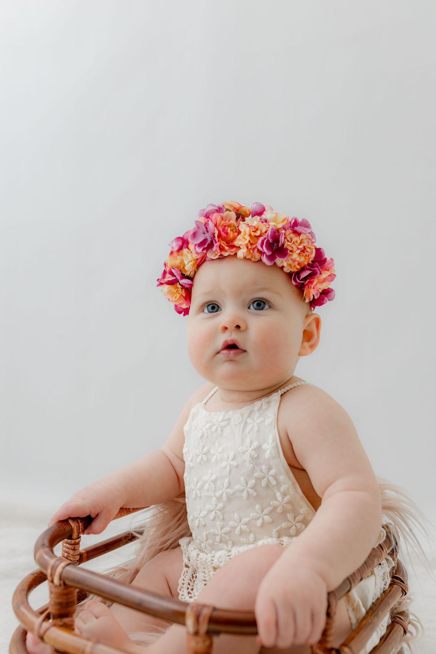 Baby girl in white lace romper wearing colorful pink and orange flower crown sitting in wicker basket.