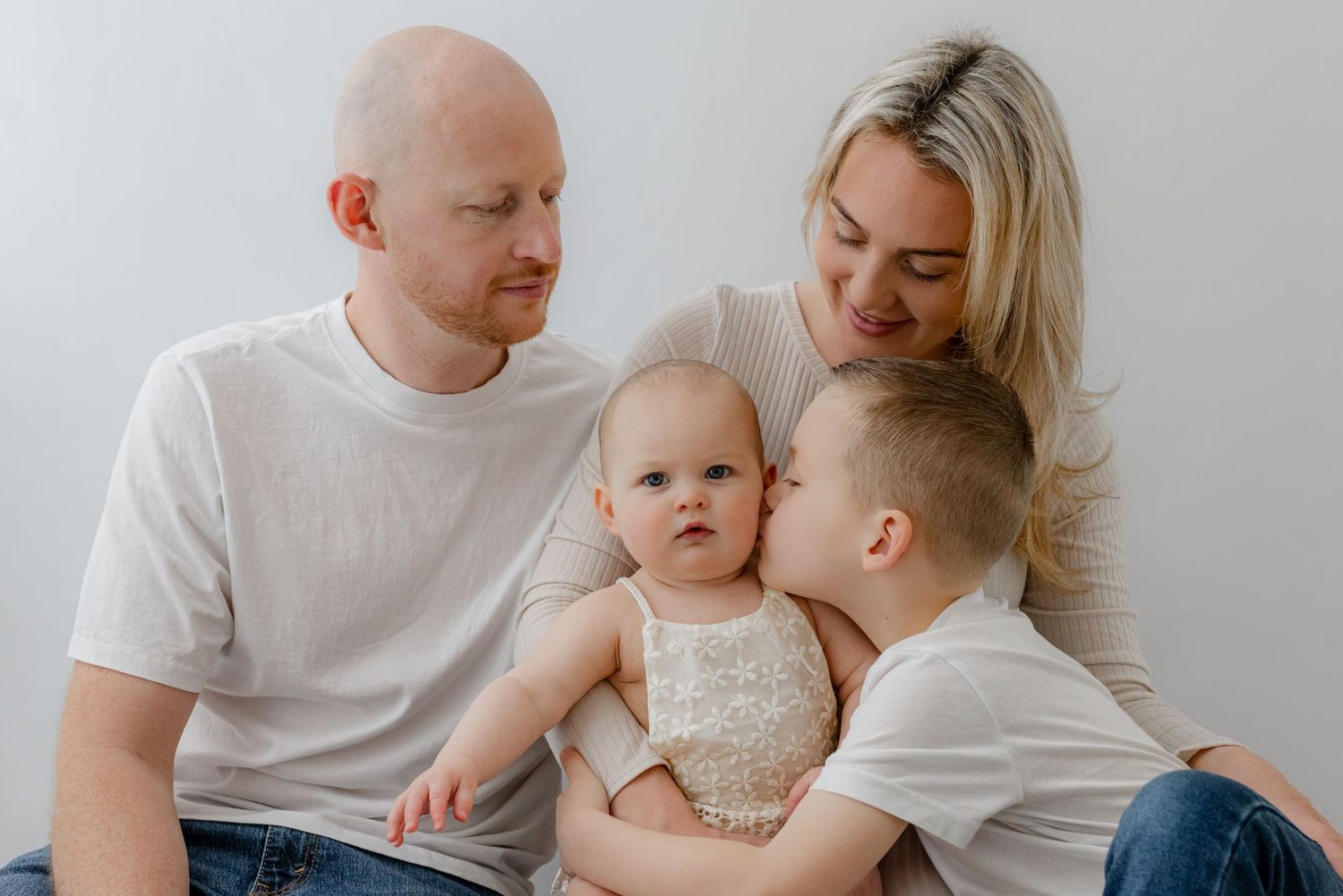 A loving family of four in white tops, parents with baby and young son sharing a tender kiss moment.