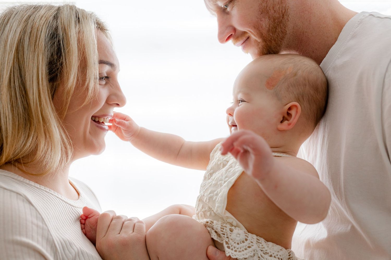 A happy young family with a smiling mother, father, and baby sharing a tender moment together in soft white light.