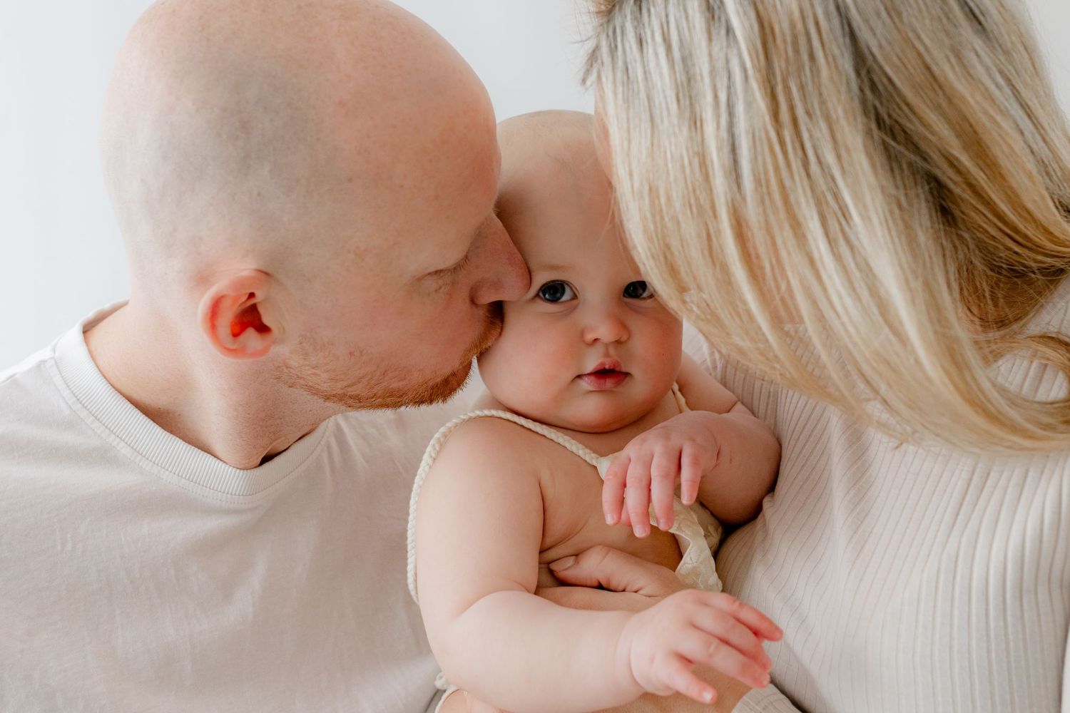 A bald father and blonde mother lovingly kiss their adorable blue-eyed baby against a white background.