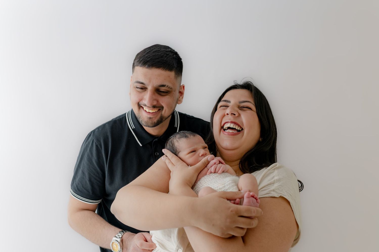 Happy couple smiling and laughing while holding their newborn baby against a white background.