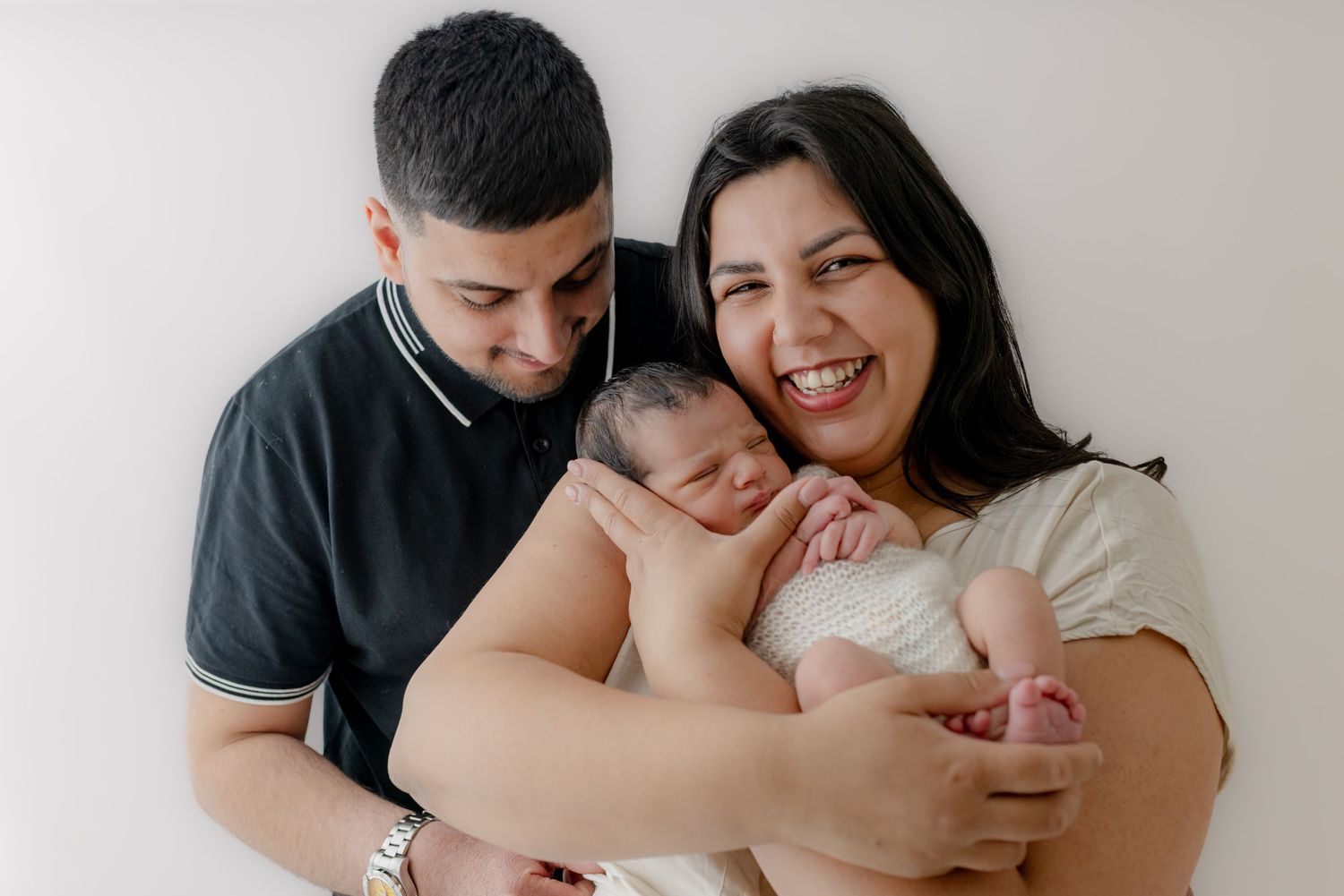 Happy parents smiling and holding their newborn baby wrapped in white against a light background.