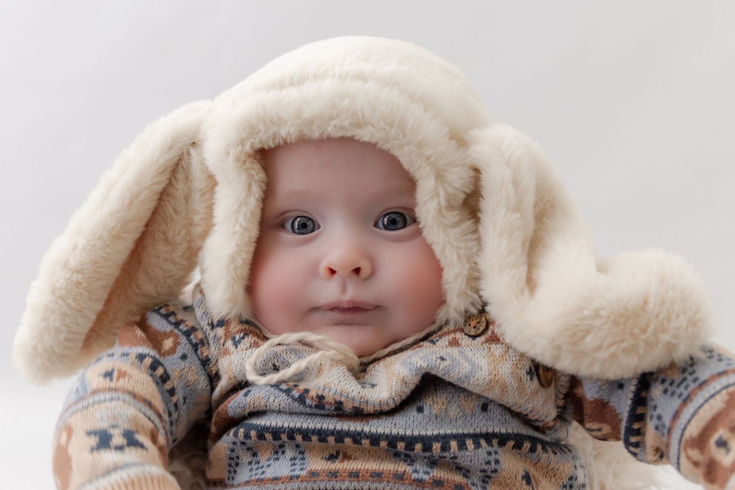 A baby wearing a cozy cream-colored bunny ear hood and patterned sweater looks directly at the camera.