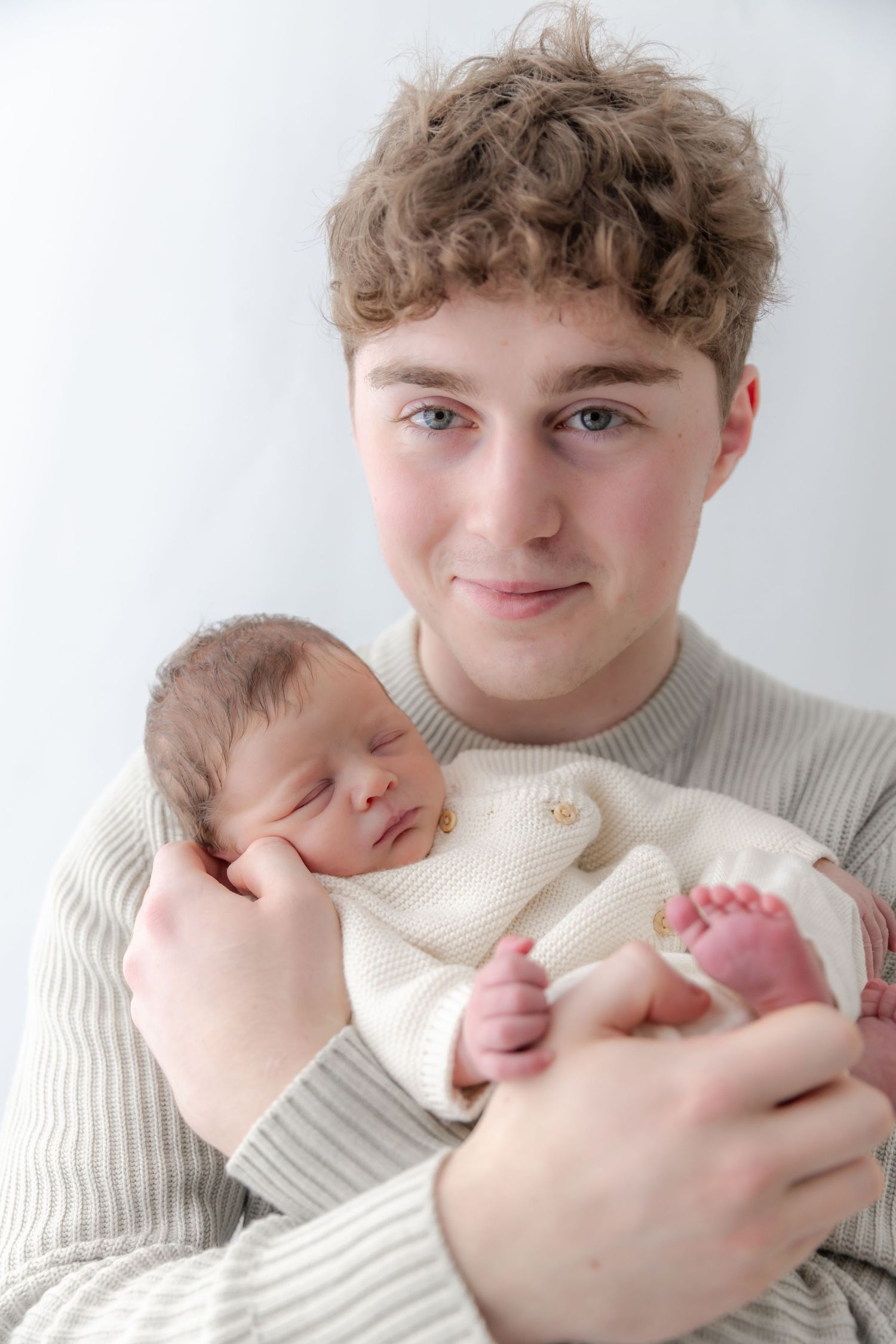 A tender moment as a young person in a cream sweater holds and gazes at a sleeping newborn baby in white clothing.