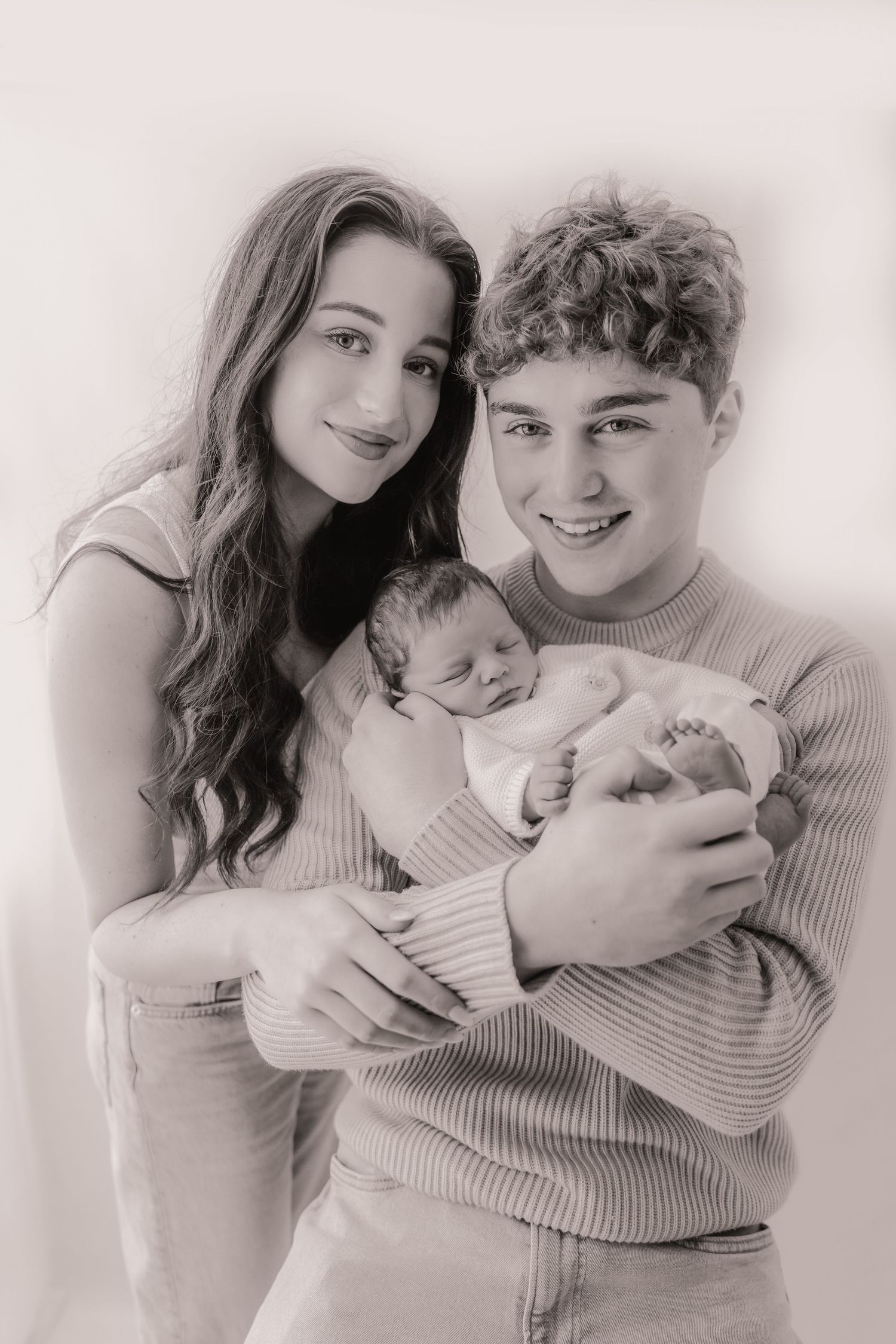 Black and white portrait of a young couple smiling and holding a newborn baby together.