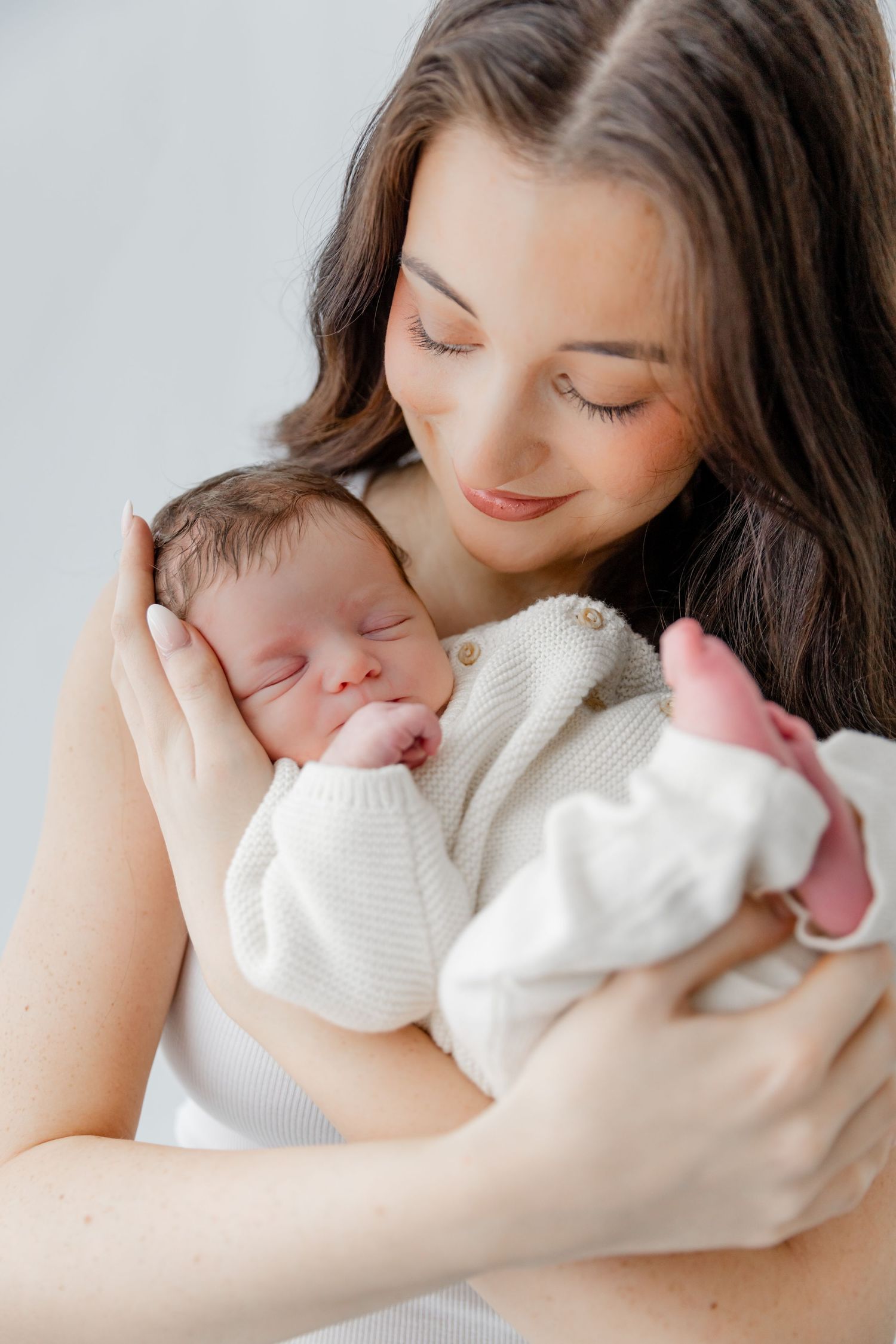 A young mother with dark hair gently cradles her sleeping newborn baby dressed in a white knit outfit.