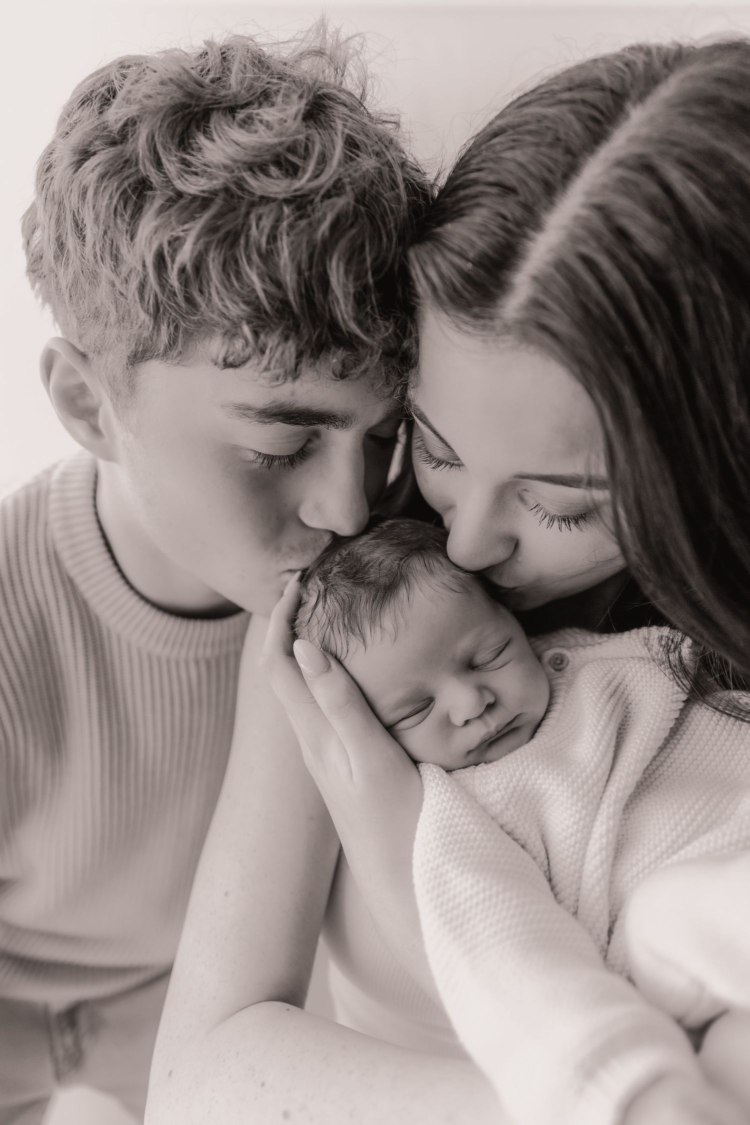 Black and white photo of two parents kissing their sleeping newborn baby, a tender family portrait.