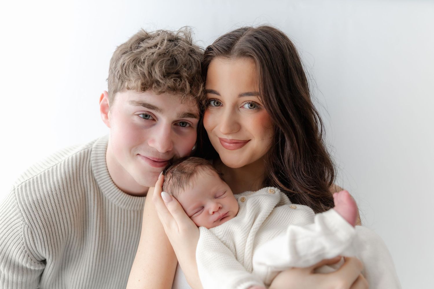 Young couple smiling and holding their sleeping newborn baby wrapped in white, posed against a white background.