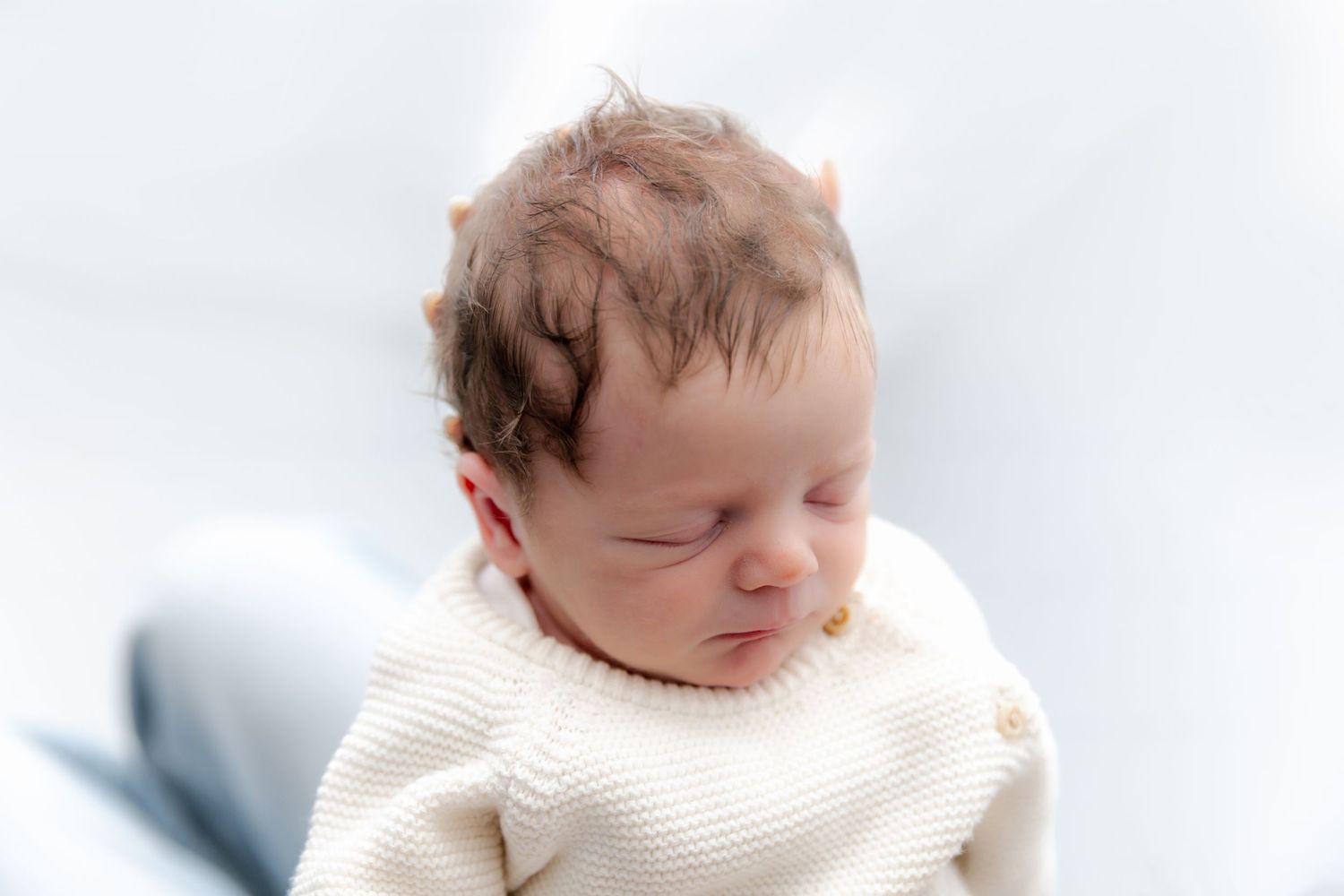 A sleeping newborn wearing a cream colored knit sweater rests against a light blue background.
