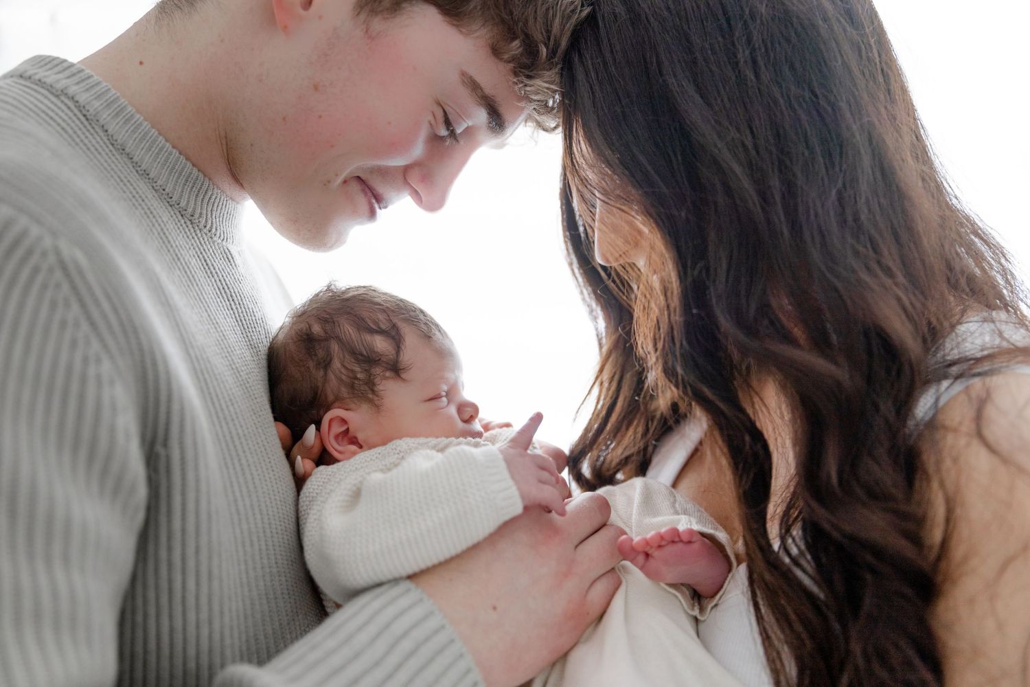 Young parents smiling lovingly at their newborn baby wrapped in white, sharing a tender family moment.