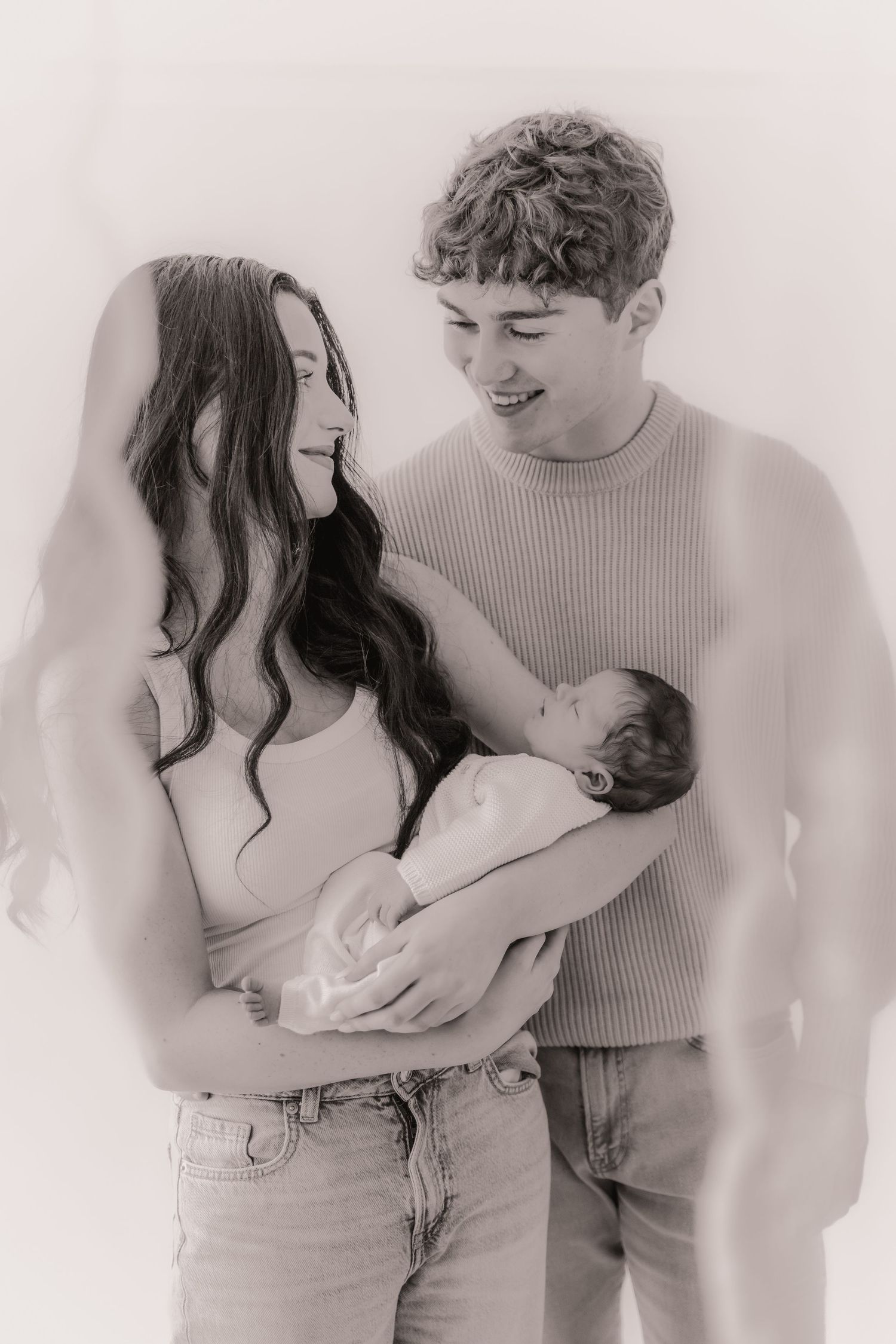 A couple in casual attire shares a tender moment with their newborn baby in a soft black and white portrait.