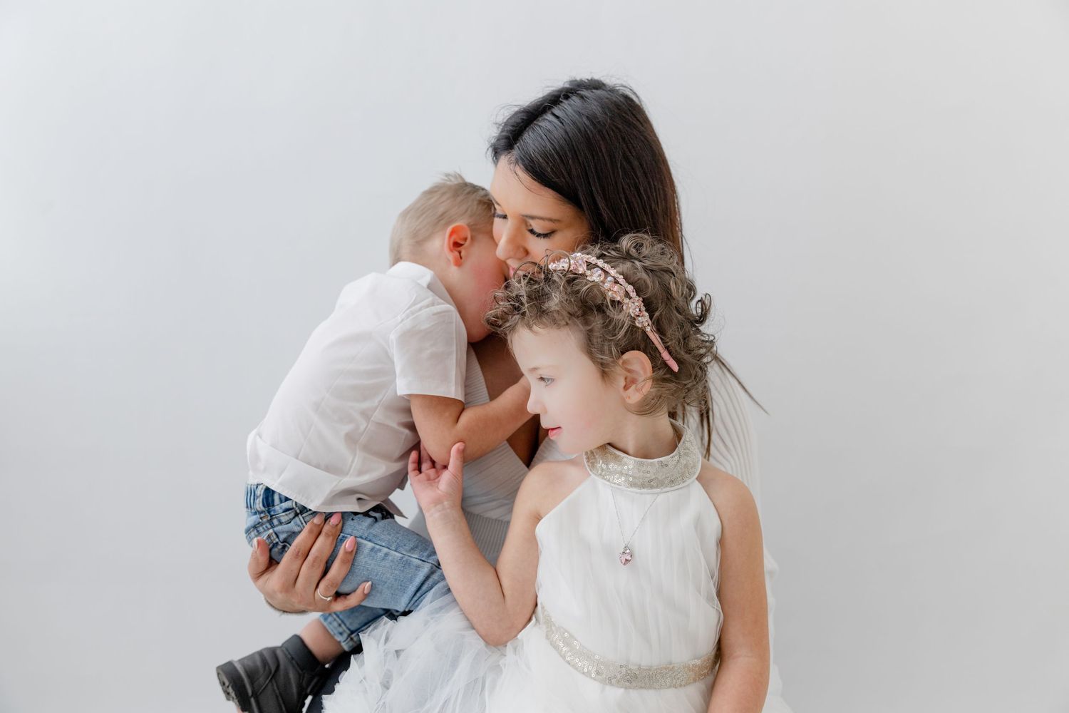 A mother holds her toddler son while her young daughter in a white dress leans in for a sweet family moment.