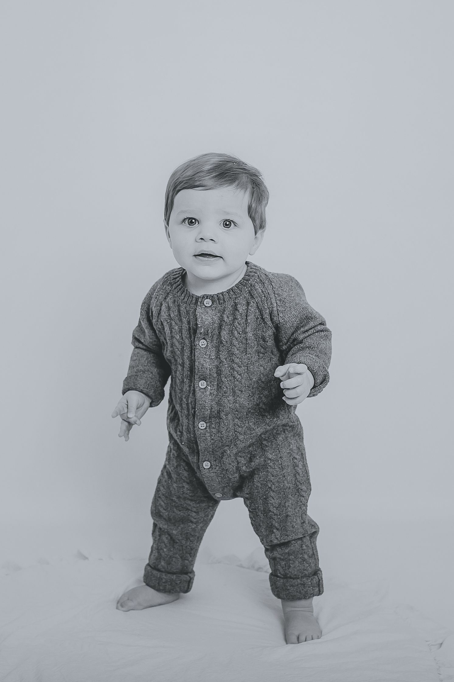 A toddler in a grey cable knit romper standing barefoot against a white backdrop. Photographer. Hull. East Yorkshire. Photography