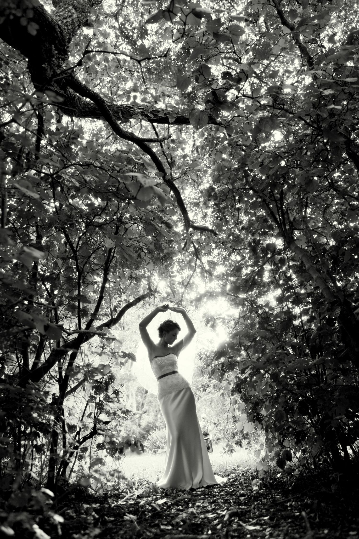 Artistic black and white photo of a figure in a flowing dress silhouetted against sunlit trees.