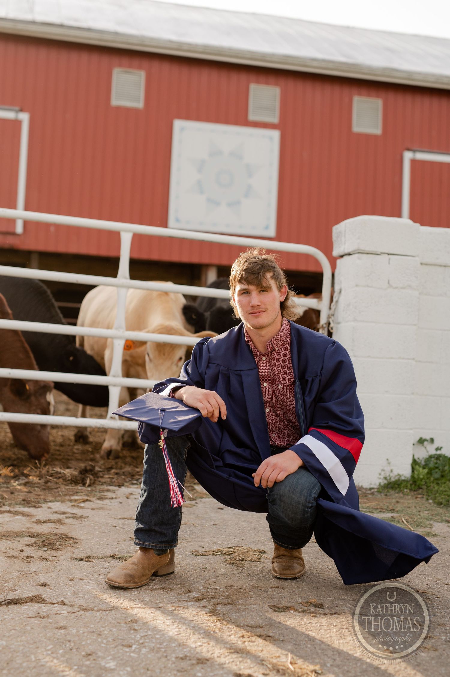 Person in graduation attire crouches near red barn with horses visible in background.